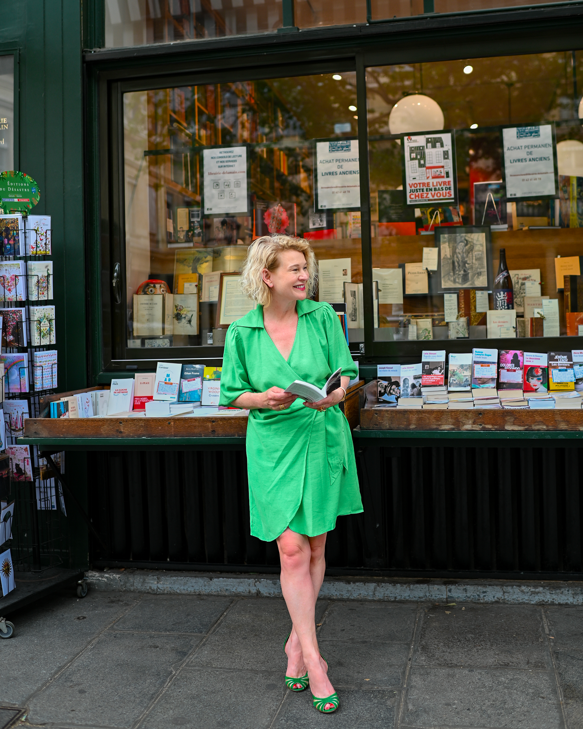 A woman with curly blonde hair, wearing a bright green dress and matching high heels, smiles while reading a book outside a bookstore with glass windows displaying books and posters.