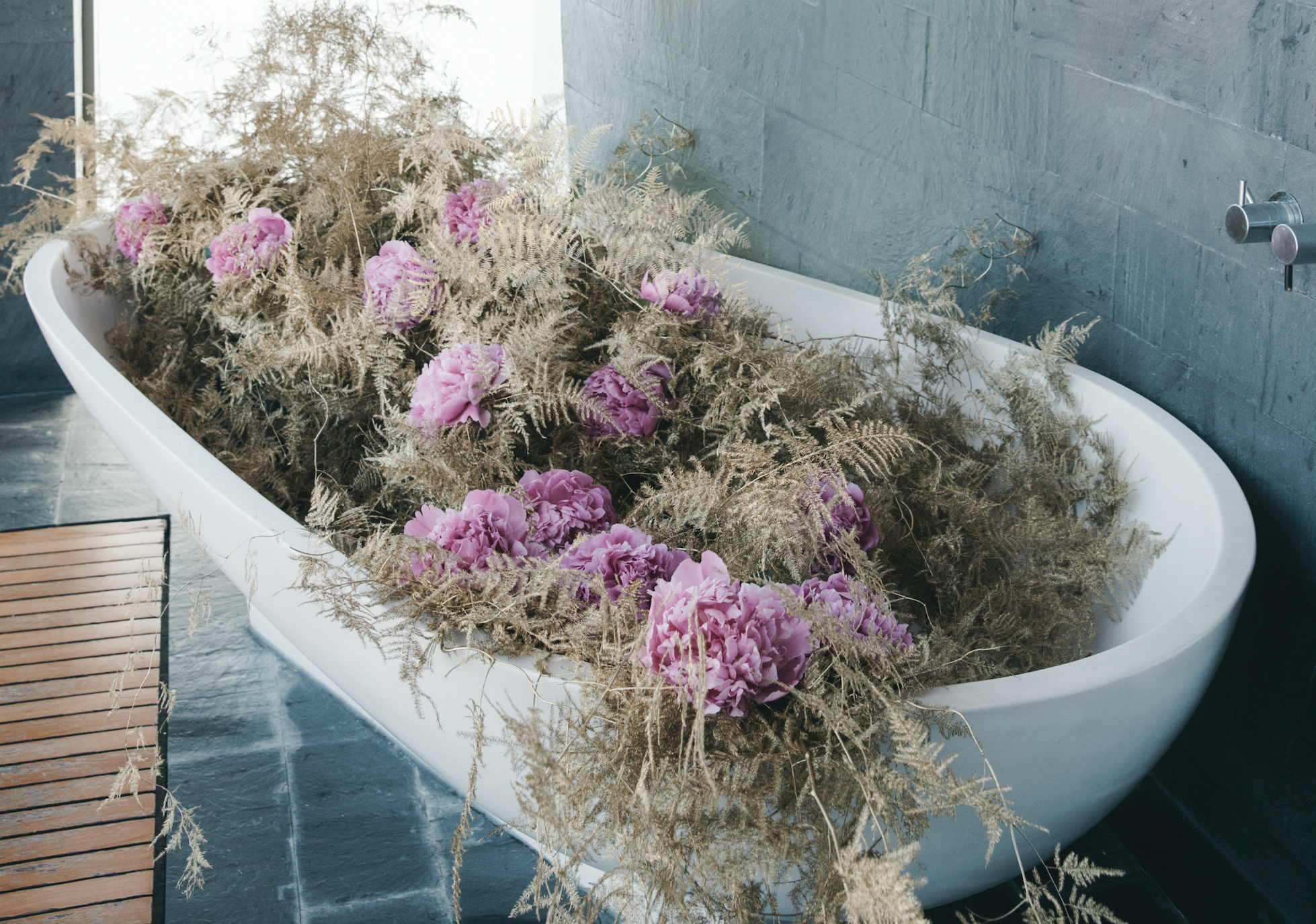 A bathroom with a white bathtub filled with dried flowers and pink peonies, gray tiled walls, a wooden bath mat, and a modern chrome faucet.