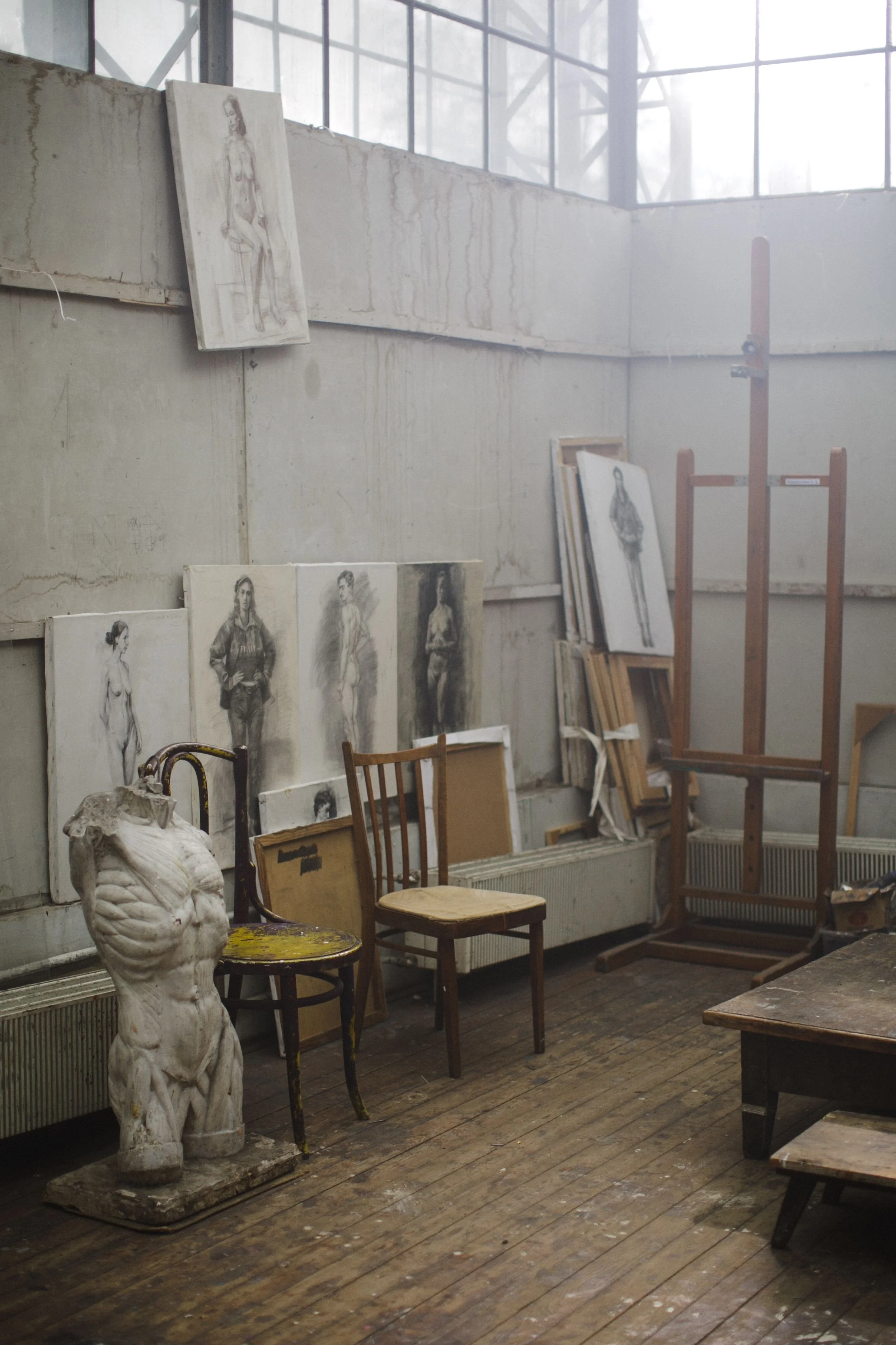 An artist's studio with sketches of female figures hanging on the wall, an unfinished sculpture of a male torso on a pedestal, and wooden chairs and tables scattered around. Large windows let in natural light.