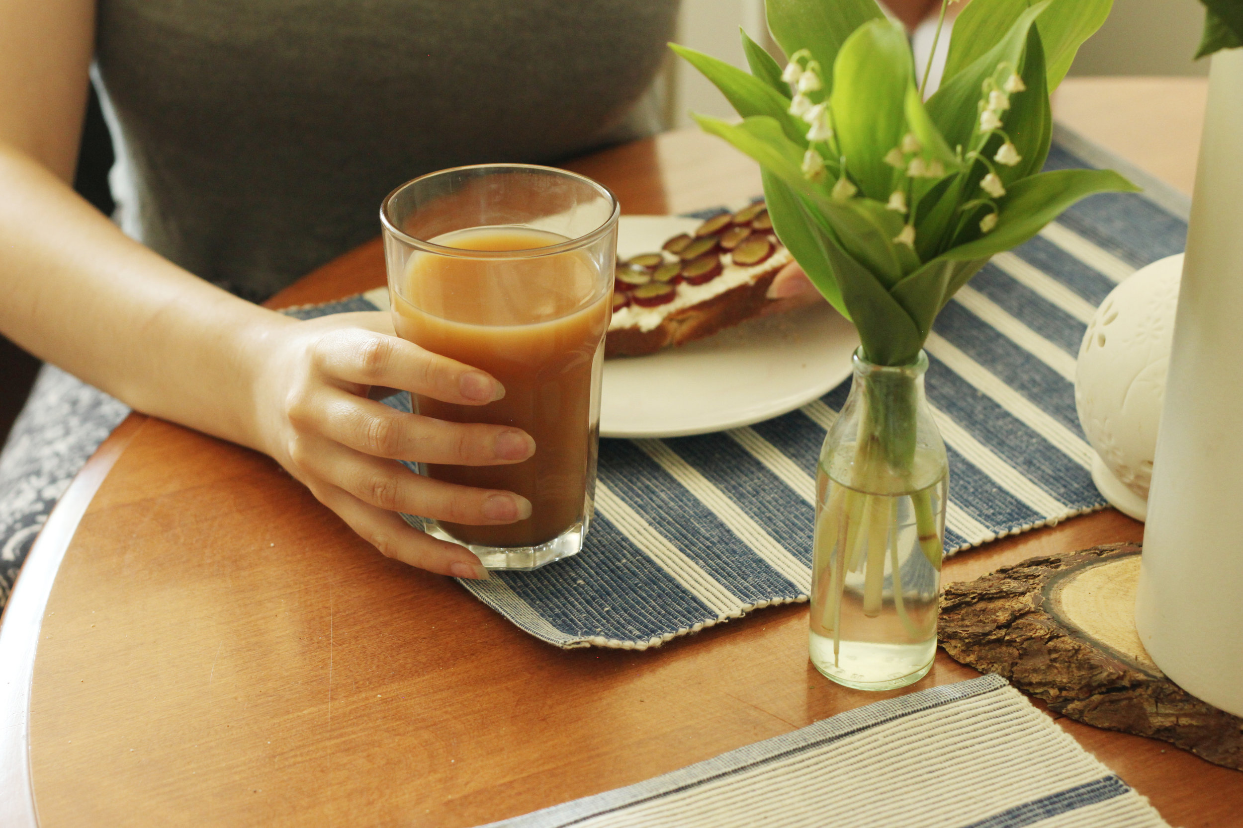 At a table, a torso down shot of someone holding a glass of coldbrew and a plate with a piece of toast.  On the table in the foreground s a small bottle with some flowers.