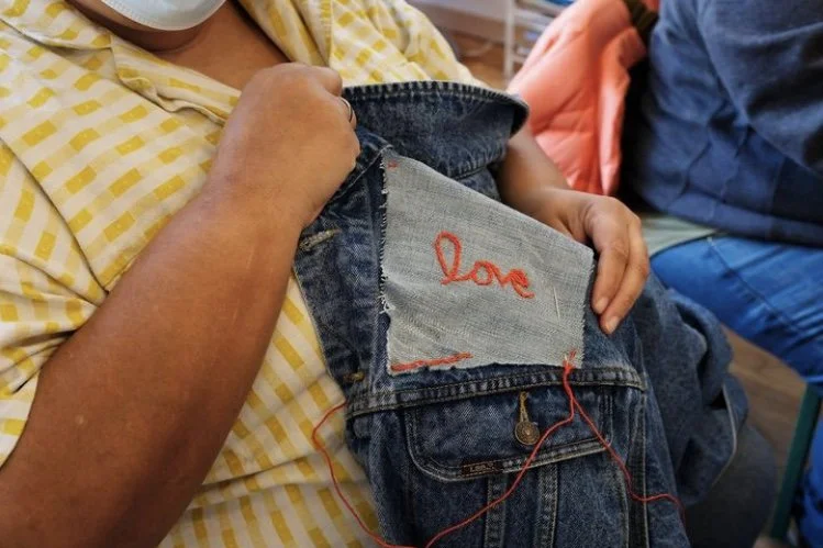 Close up shot of person's hands holding a denim jacket with an embroidered patch reading 'love' in red cursive stitching.