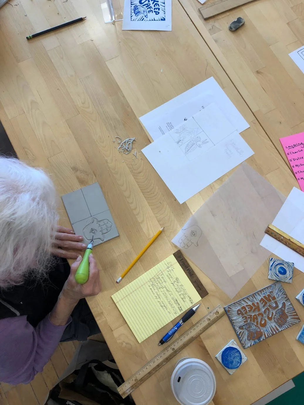 Overhead shot of participant carving a linoleum block. Shavings, sketches, and notes visible on wooden table.