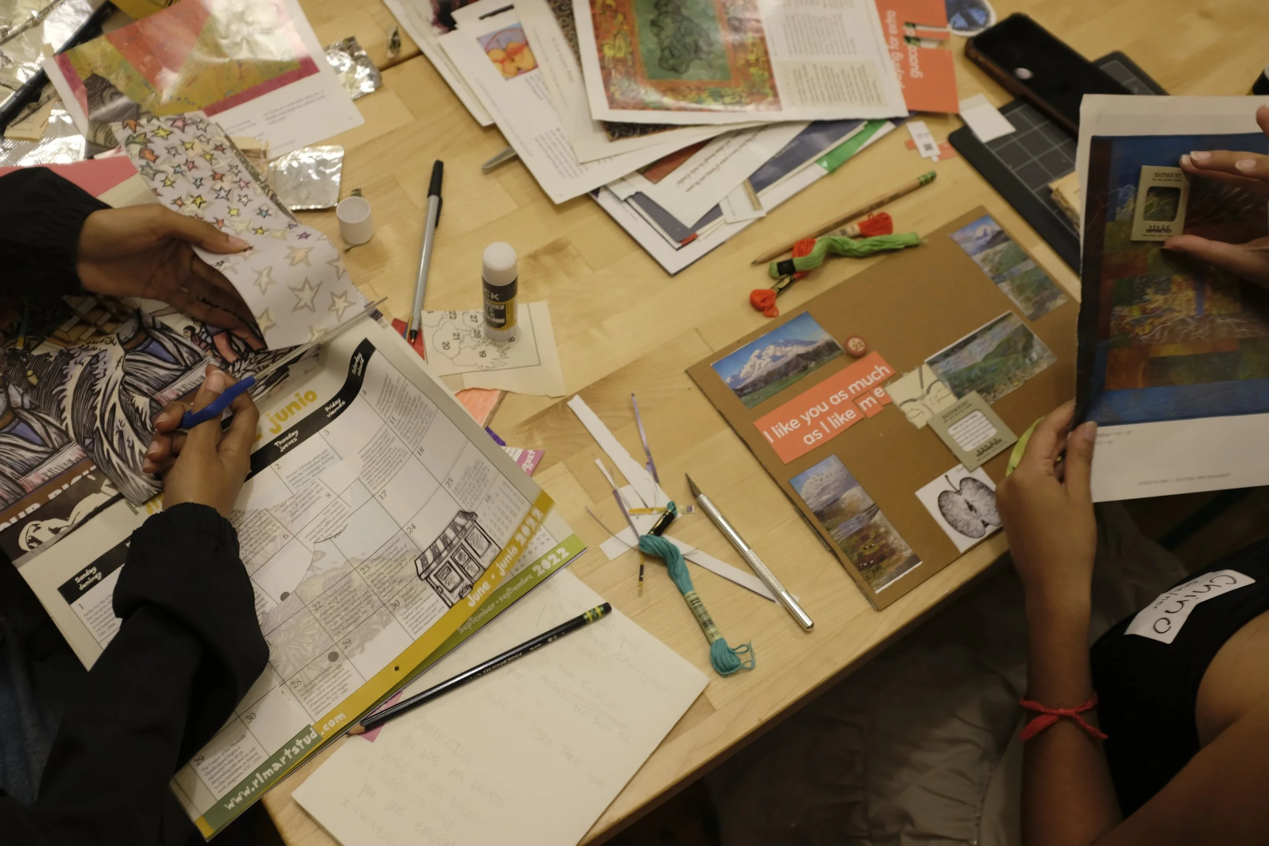 Overhead photo of two people collaging. Collage scraps and in progress collages are scattered on the table.