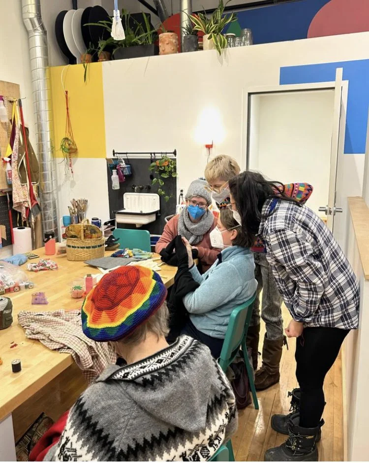 Group of 4 participants huddle standing around one seated participant doing a mending demonstration. Mending supplies are scattered on table in front of them.