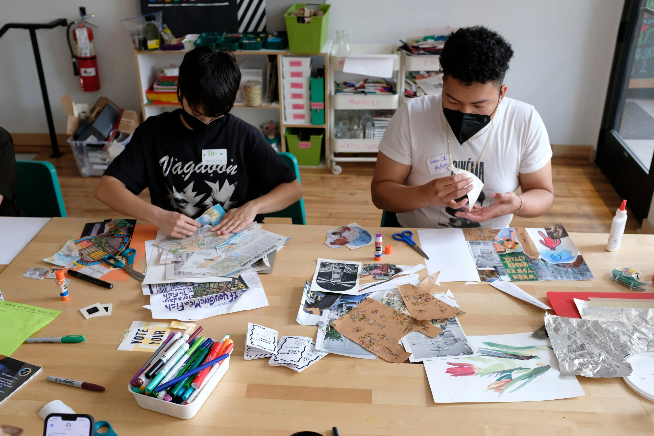 Front shot of two masked participants sitting at table collaging. Collage materials are scattered on the table in front of them.