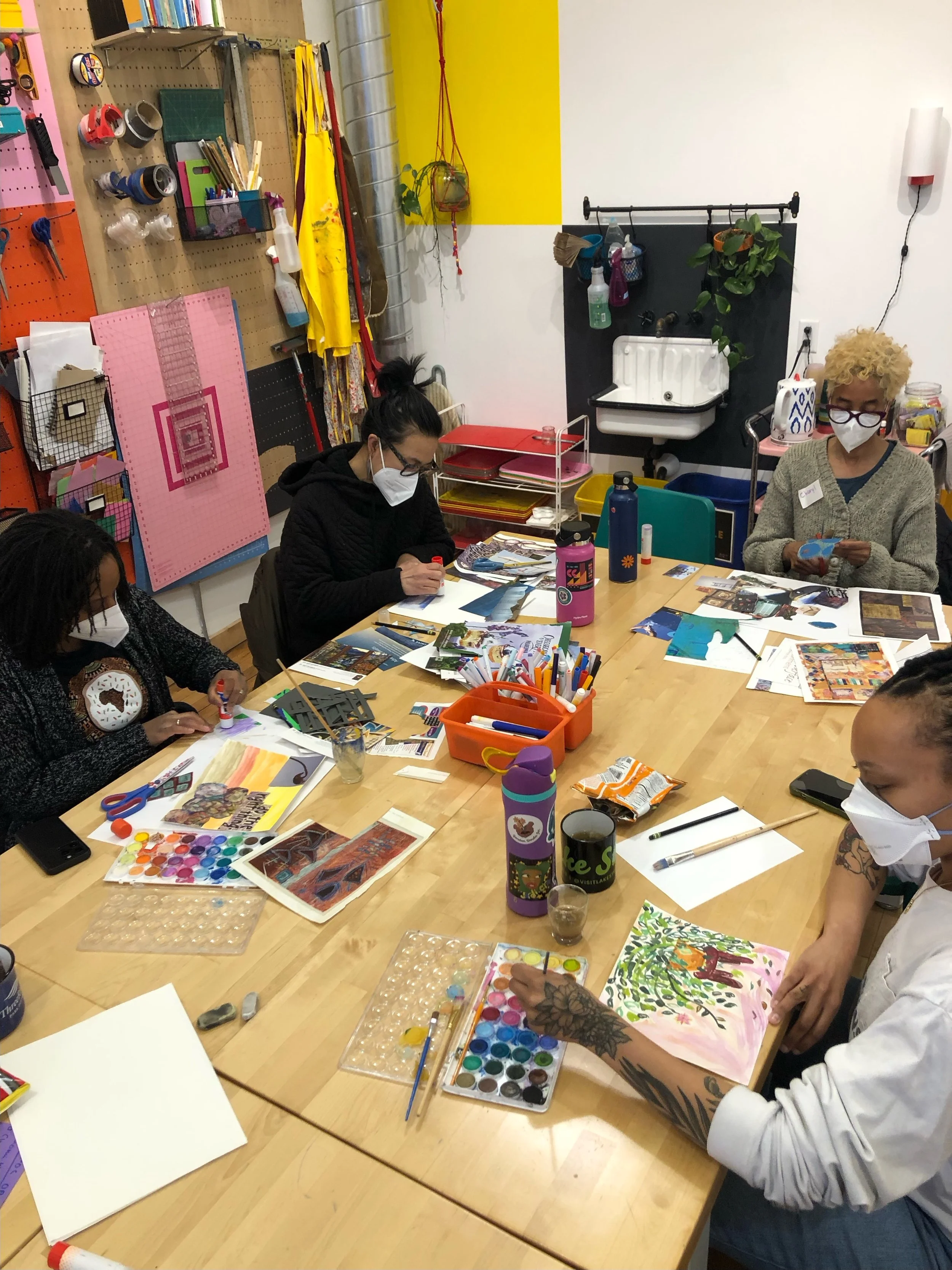 Group shot of 4 masked participants sitting at a table. Some are painting with watercolor and others are collaging. Art materials scatter the table.