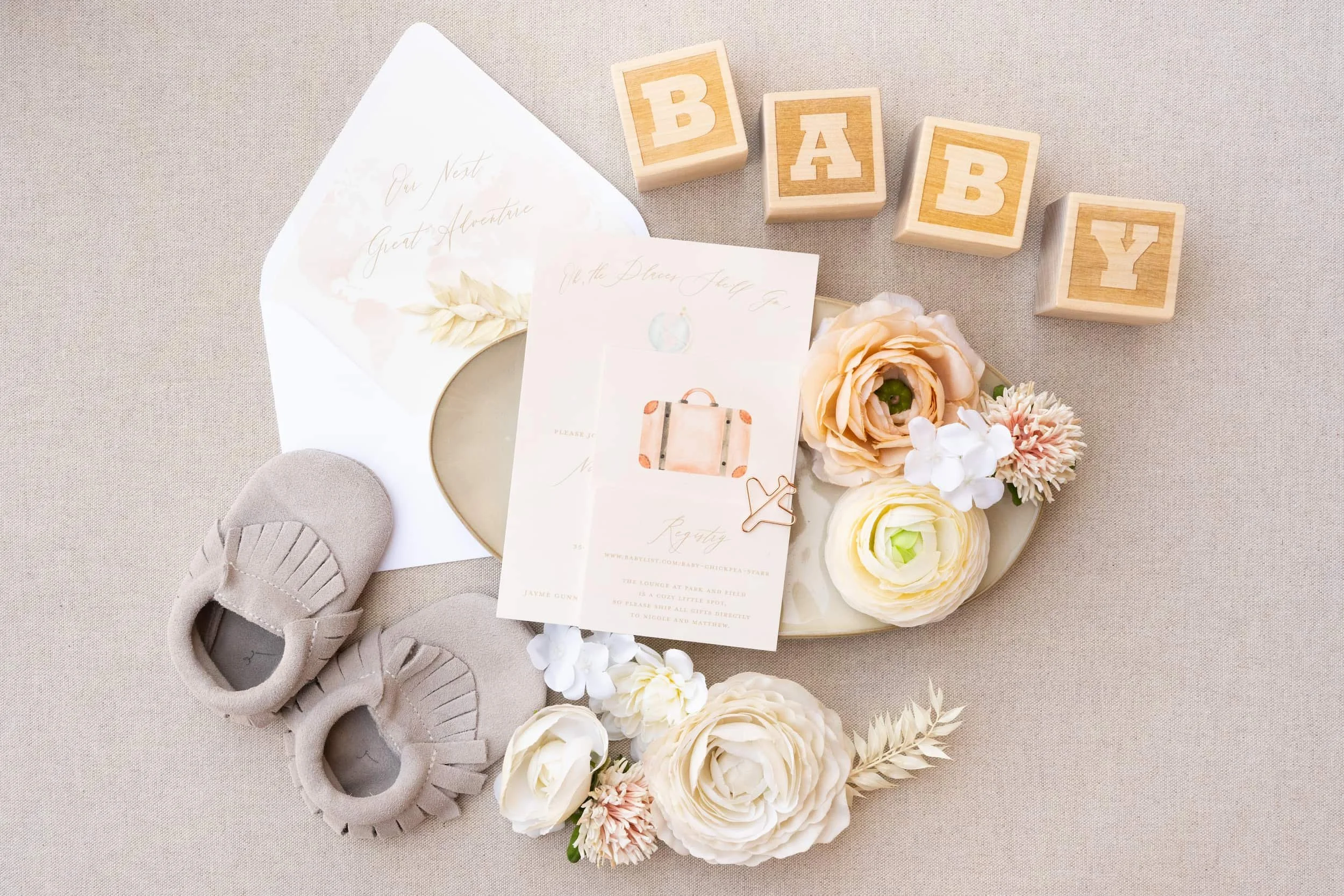 Flat lay of an adventure-themed baby shower invitation suite featuring peach and sage watercolor artwork, airplane paper clips, and travel-inspired stationery for a couple celebrating their next great adventure—welcoming a new baby.