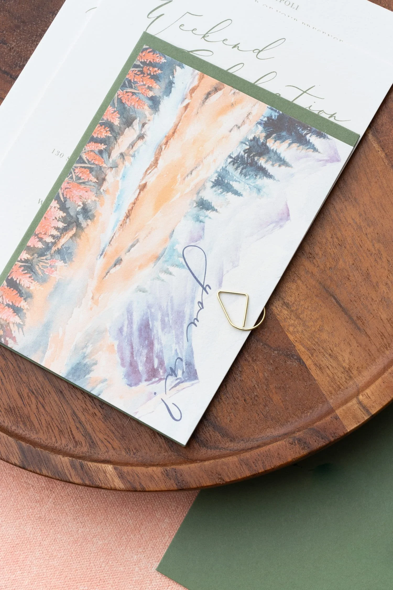 Luxury mountain ranch wedding invitation suite featuring watercolor autumn mountain landscapes, peach and sage color palette, modern script typography, handmade paper, and Western-inspired details arranged in an elegant flat lay.