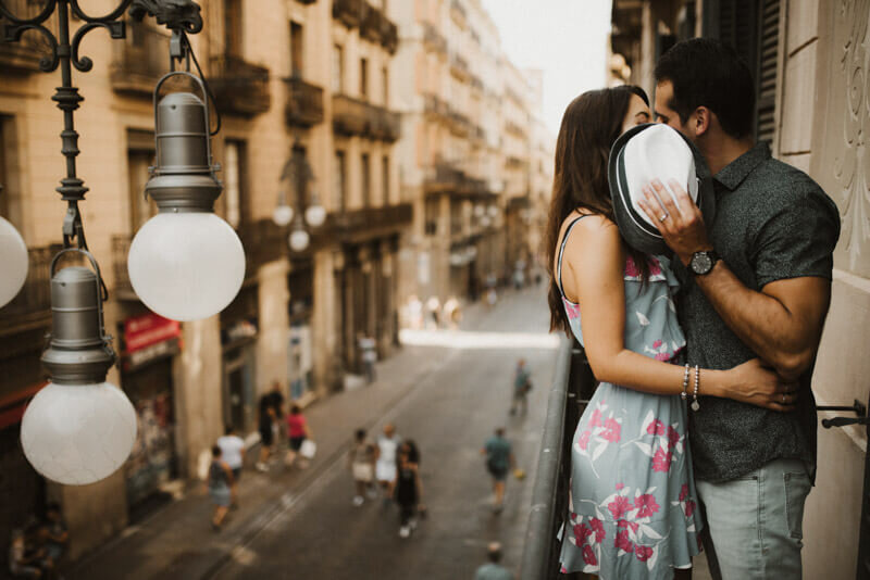 Couple posing gothic quarter balcony hiding kiss with hat