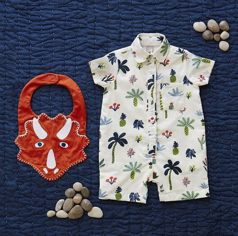 Children's outfit with a white shirt featuring a tropical pattern of palms, pineapples, and cacti, laid out on a dark blue quilted blanket. Accompanying the shirt are a red and white animal-shaped bib or accessory and small rocks arranged nearby.