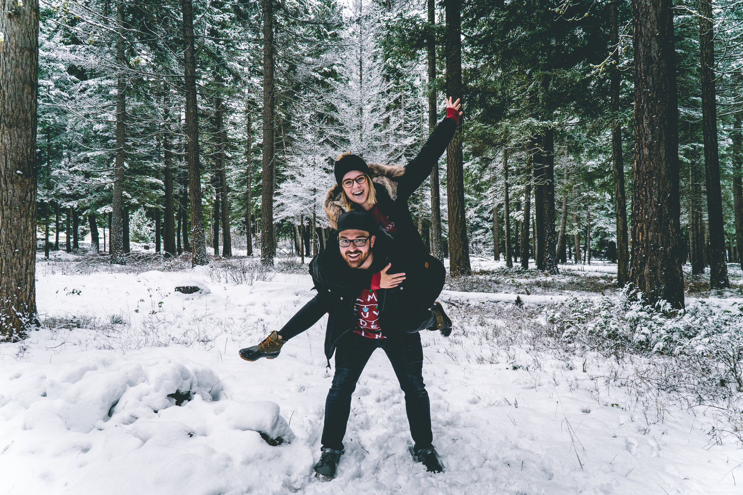 Two people, a man and a woman, are in a snowy forest in Cle Elum Washington, with the woman on the man's shoulders, smiling and waving, while the man is holding her legs.