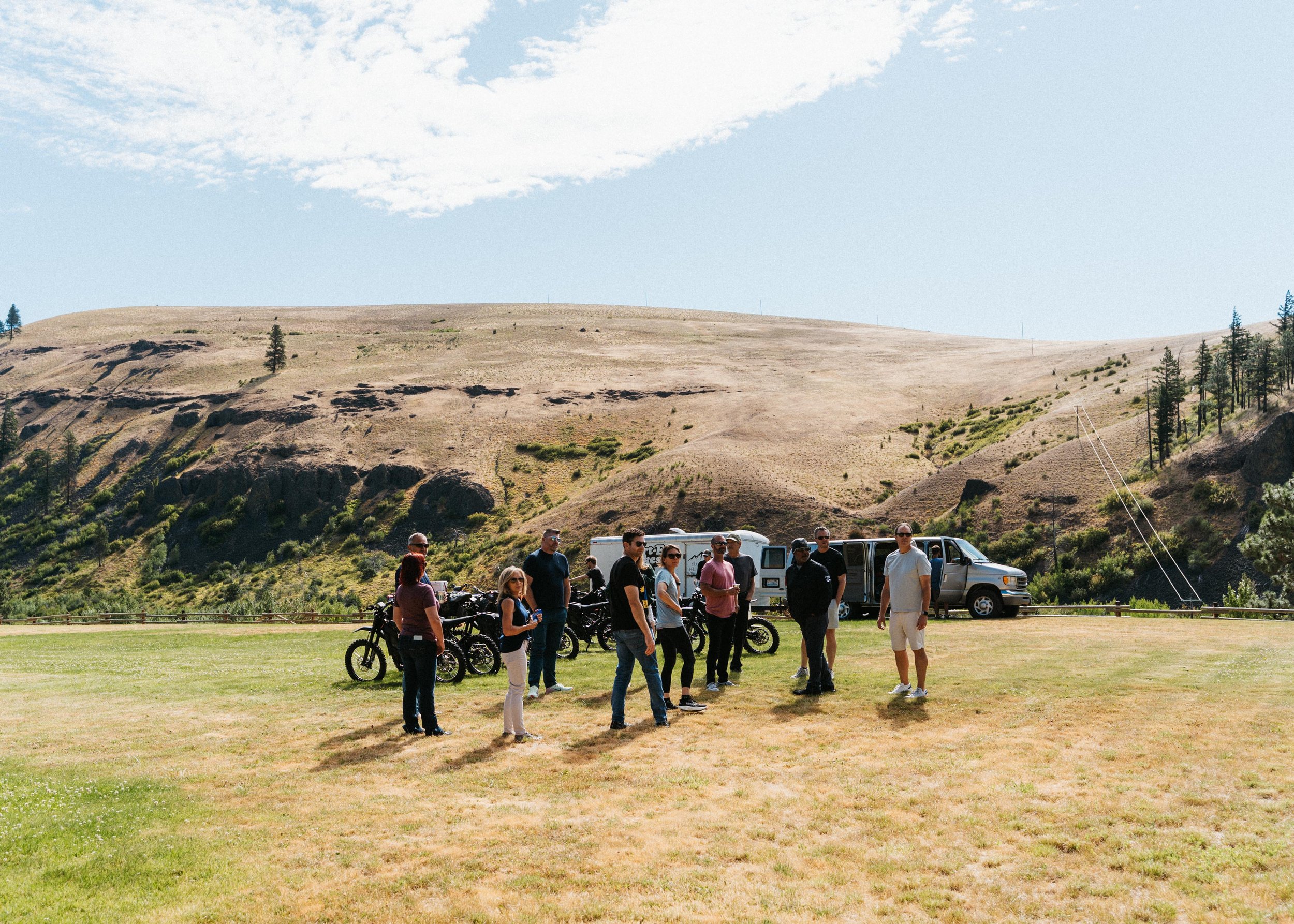 Group of people with motorcycles and vans gathered in a grassy mountain landscape beneath a partly cloudy sky