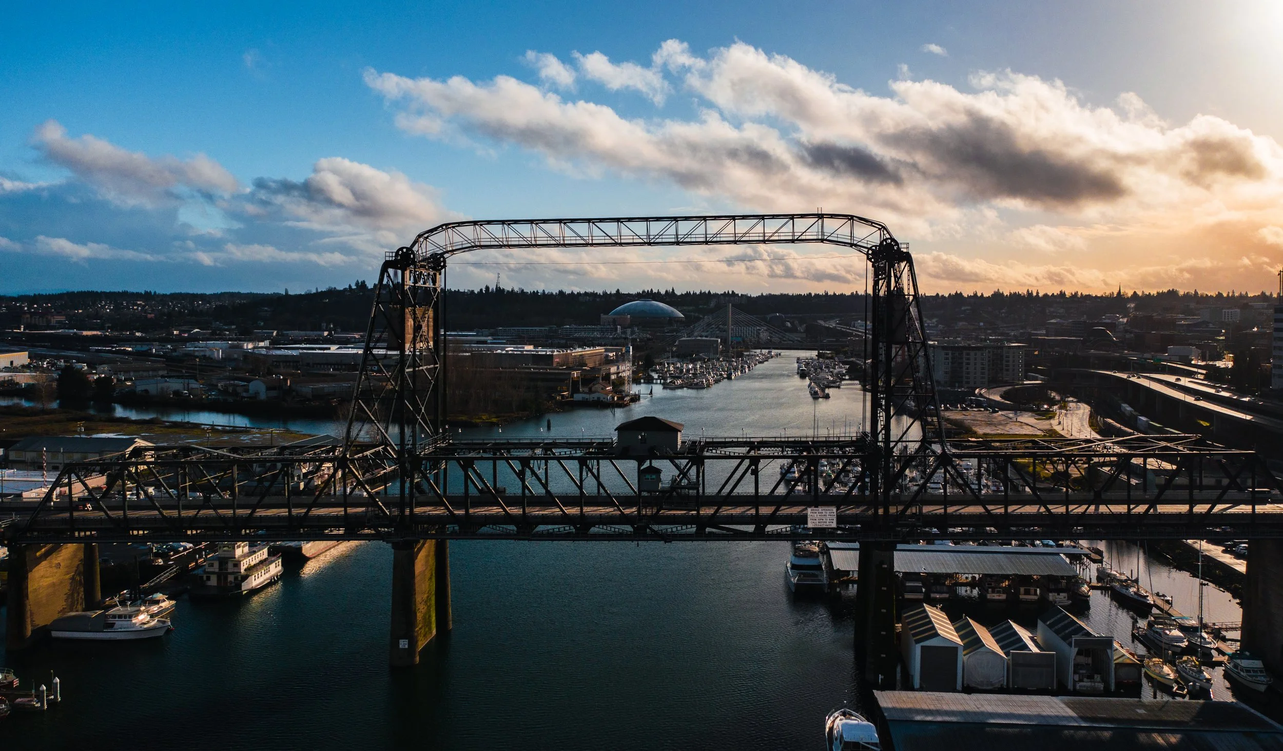 View of a city with a waterway, boats, and a bridge during sunset, with cloudy sky.