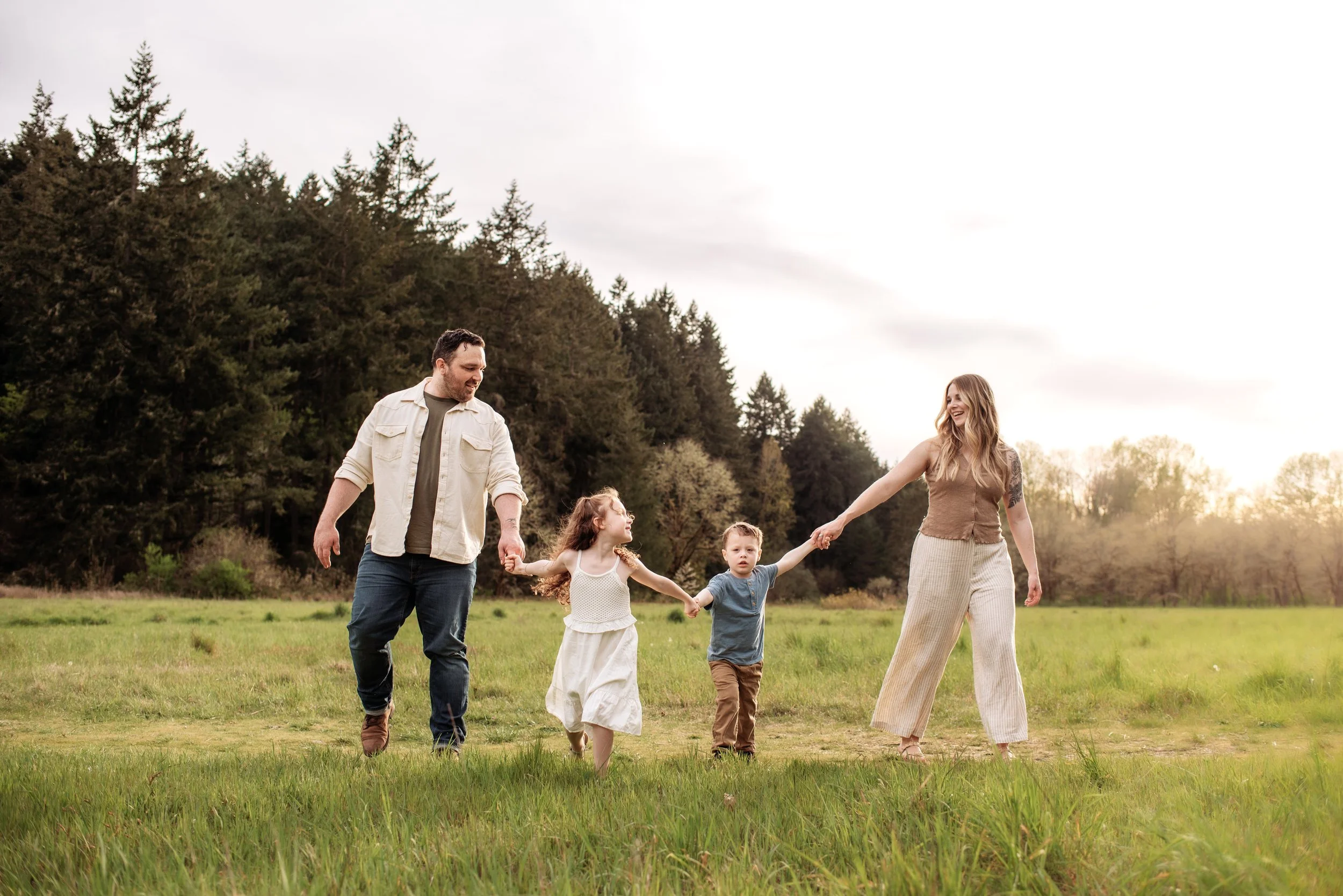 A family of four, including two adults and two children, walking hand-in-hand through a grassy field with trees and a cloudy sky in the background in fort steilacoom park