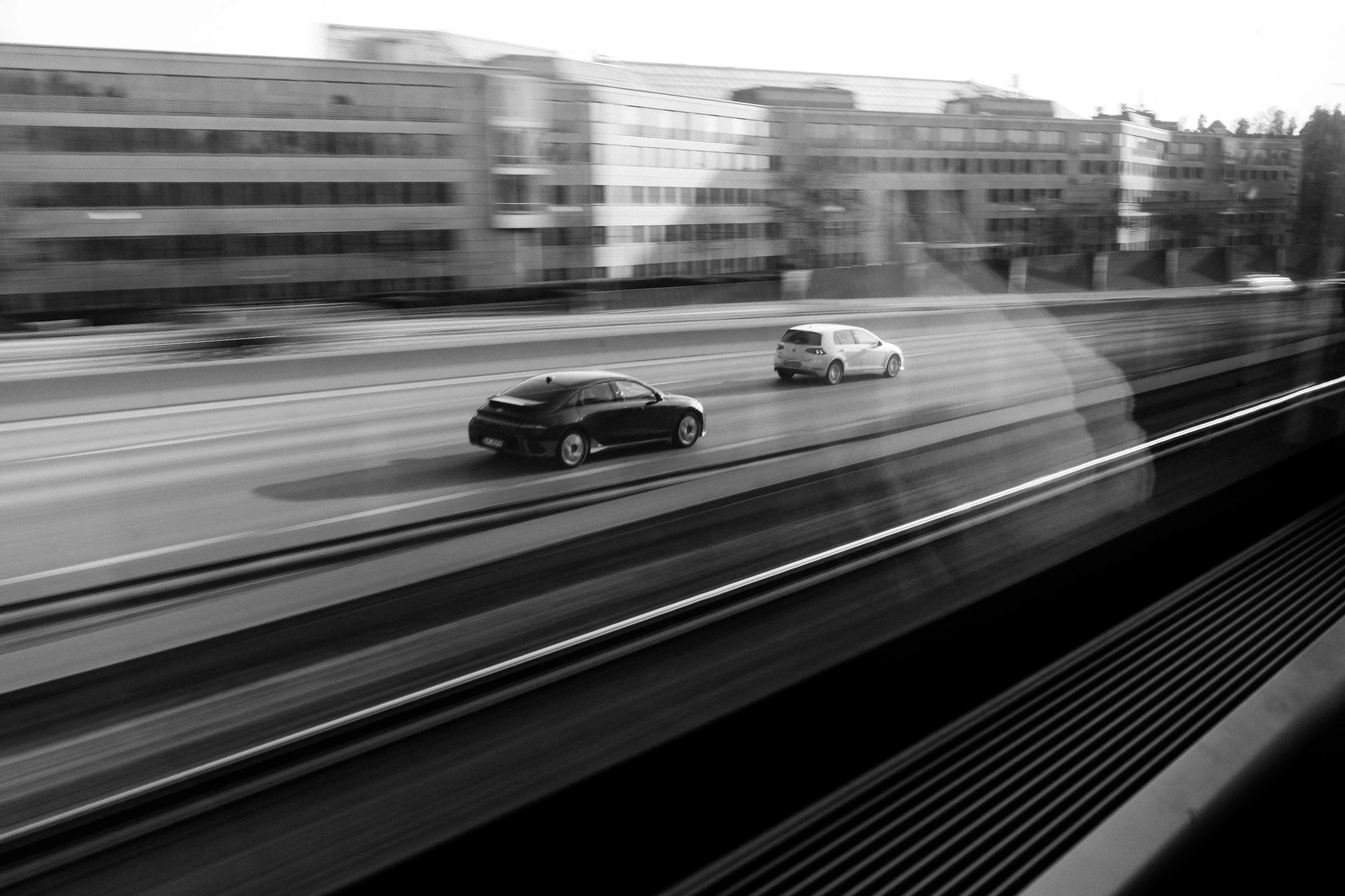 Black and white photo of moving cars on a highway viewed from a train window with blurred lines indicating motion, and a cityscape with buildings in the background.