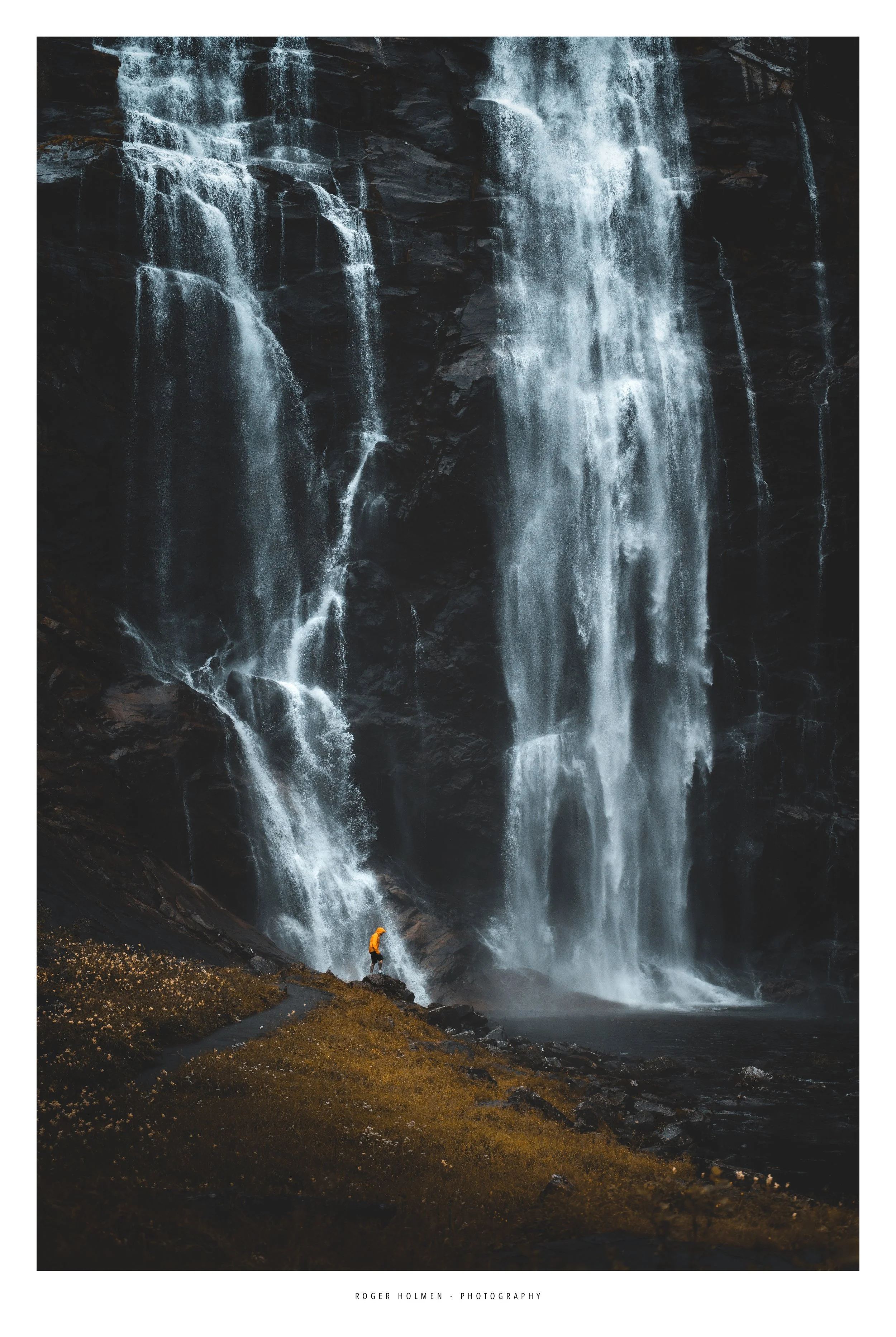 A person in a bright orange jacket standing on the ground near a waterfall, with large streams of water cascading down a dark rock face.