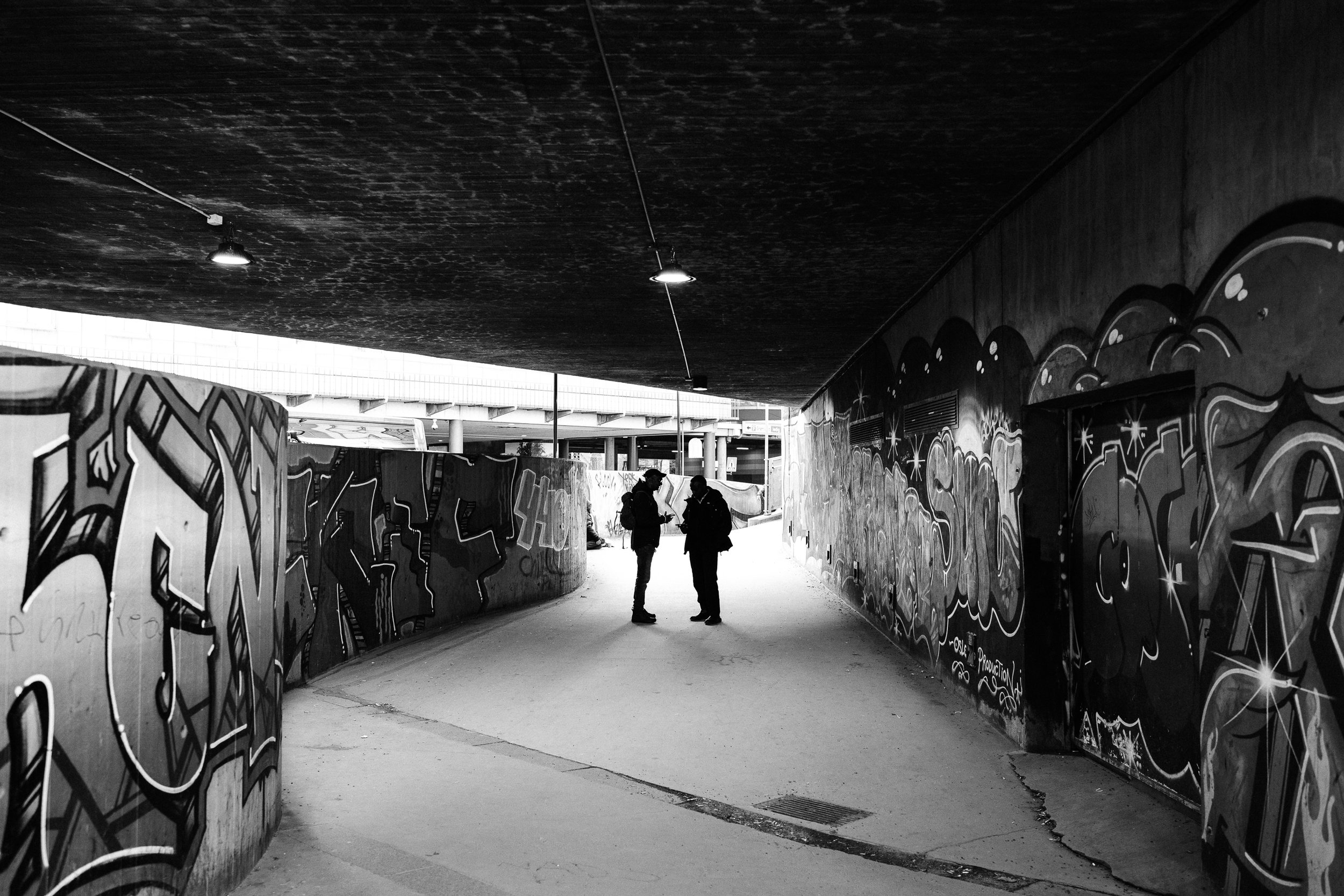 A black and white photo of a tunnel with graffiti on the walls, two people standing and talking in the middle, with a bright outdoor area in the background.