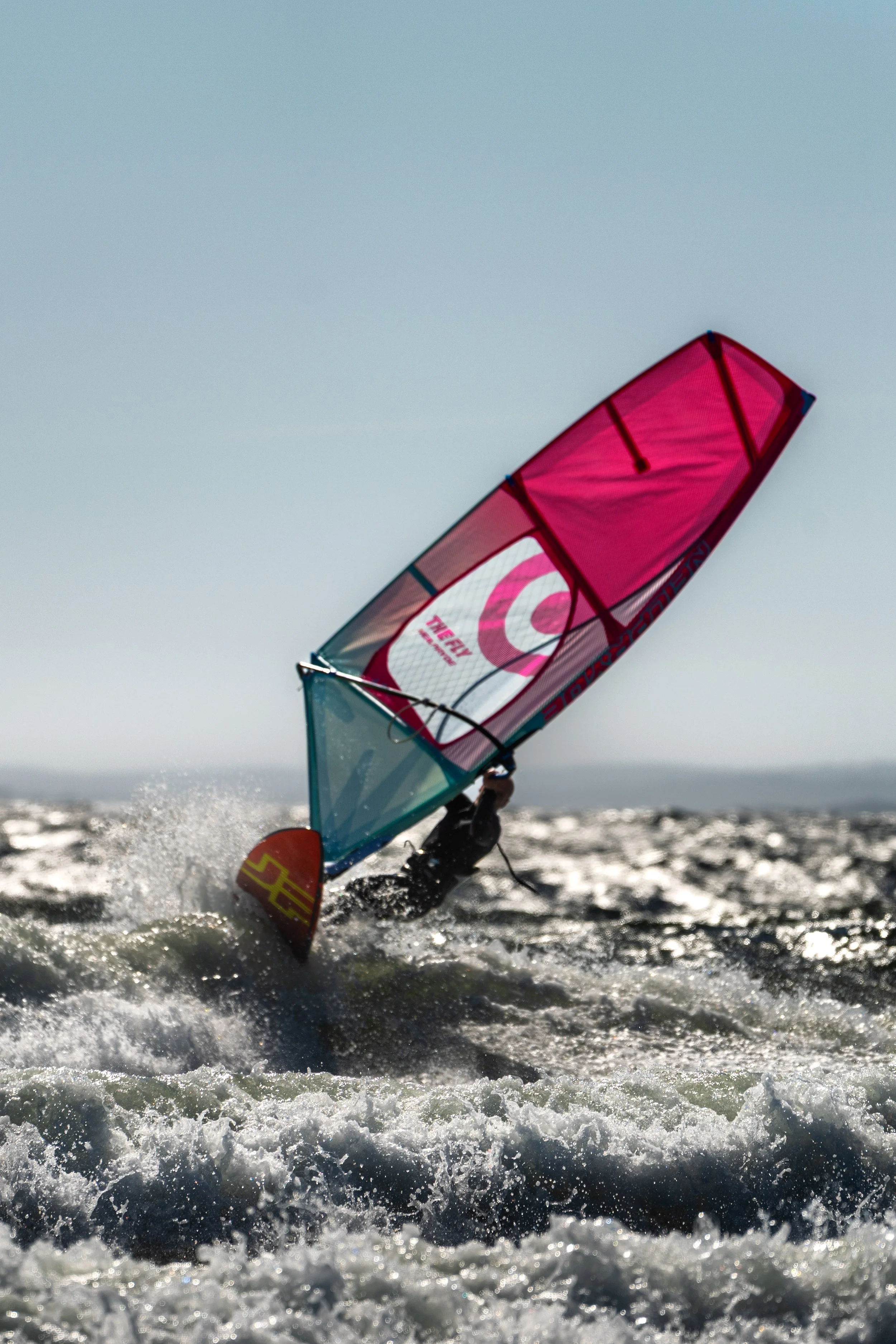A person windsurfing on the water with a pink sail, kicking up water spray in the sunlight.
