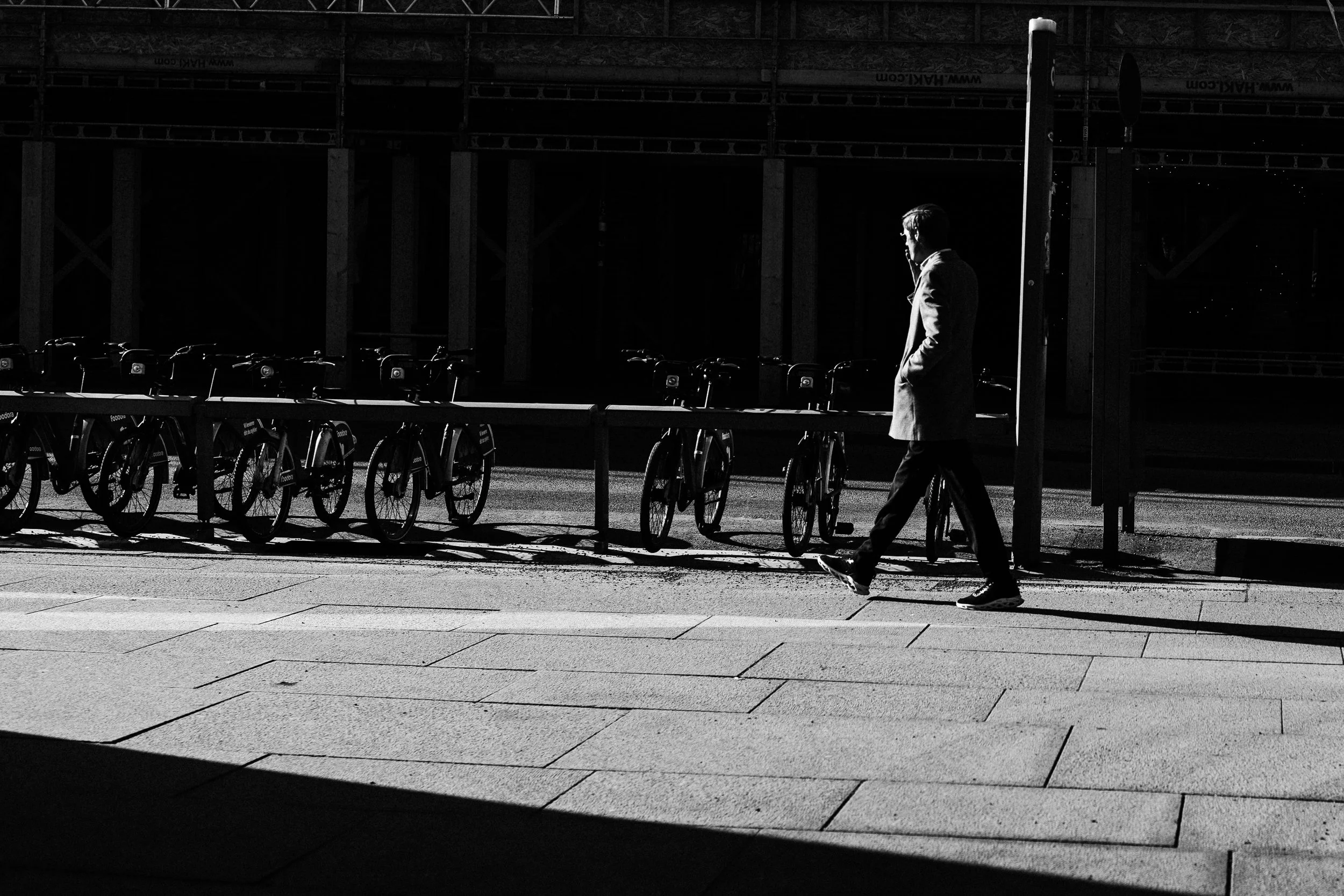 A man walking on a city sidewalk near a row of parked bicycles, with a shadowed building in the background in black and white.