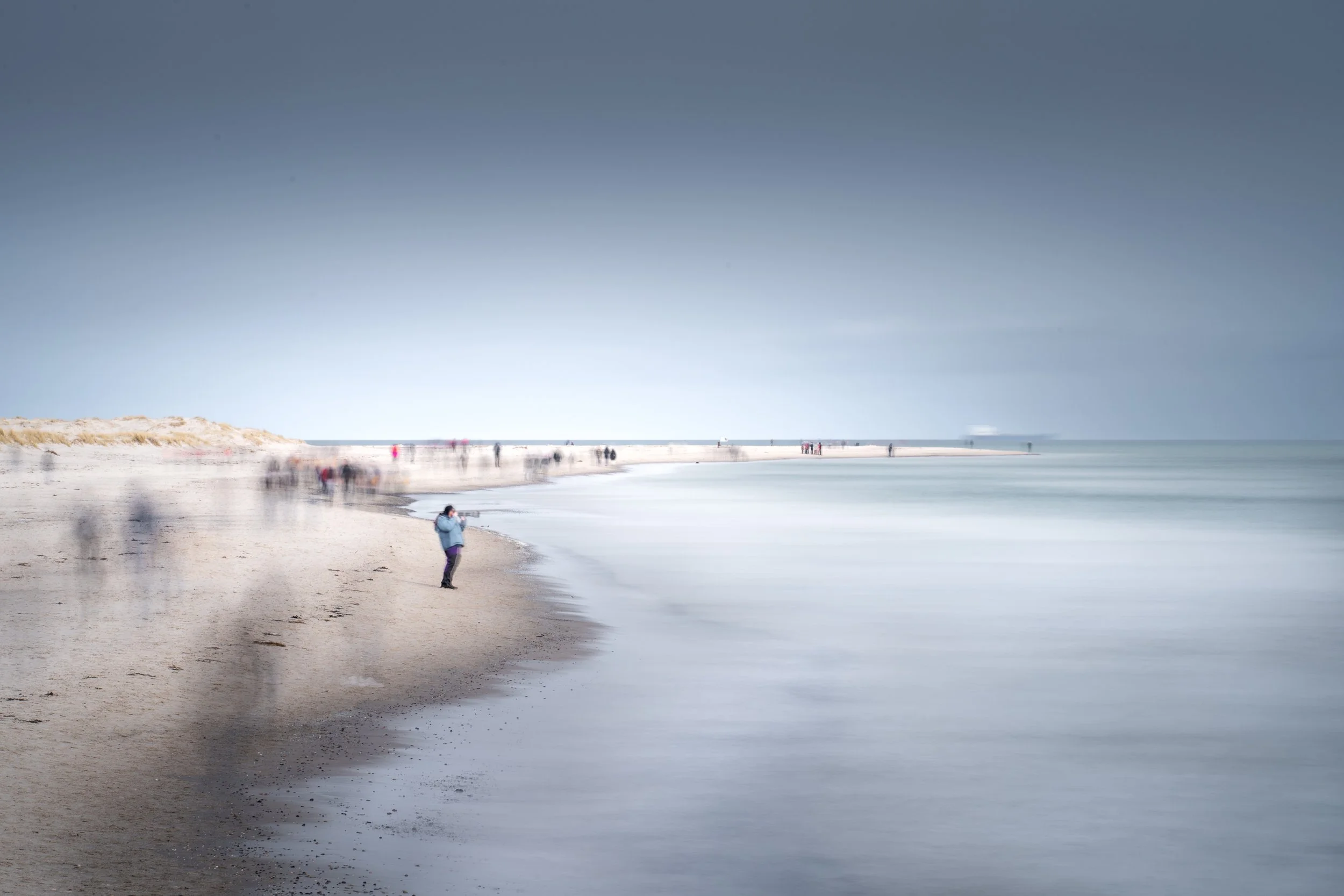 A long sandy beach with a person taking a photo near the shoreline. There are several blurred figures walking along the beach and a pier extending into the water. The sky is cloudy and the photo has a slow shutter effect creating a smooth, blurred mo