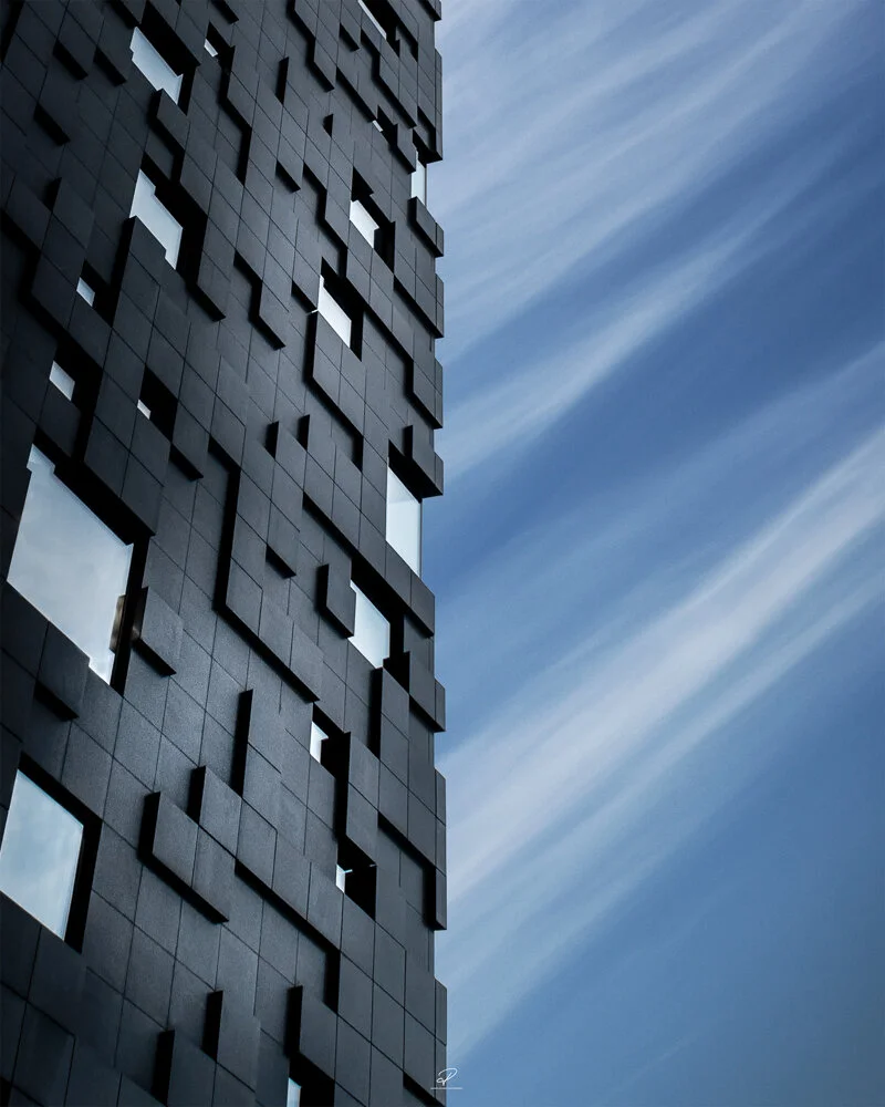 A tall modern building with a geometric facade consisting of black square panels and window openings, set against a blue sky with streaks of white clouds. Arkitektur foto Oslo.