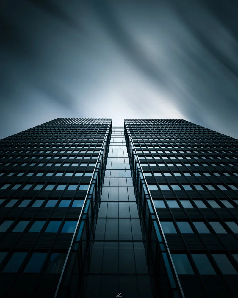 A low-angle view of a tall, modern glass office building with a cloudy sky in the background. Arkitektur foto Oslo.