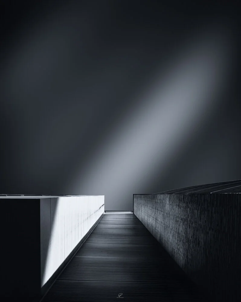 A minimalist black and white photograph of a bridge or walkway with a high wall on the right and a low wall on the left, leading towards a foggy or cloudy sky. Arkitektur foto Oslo.