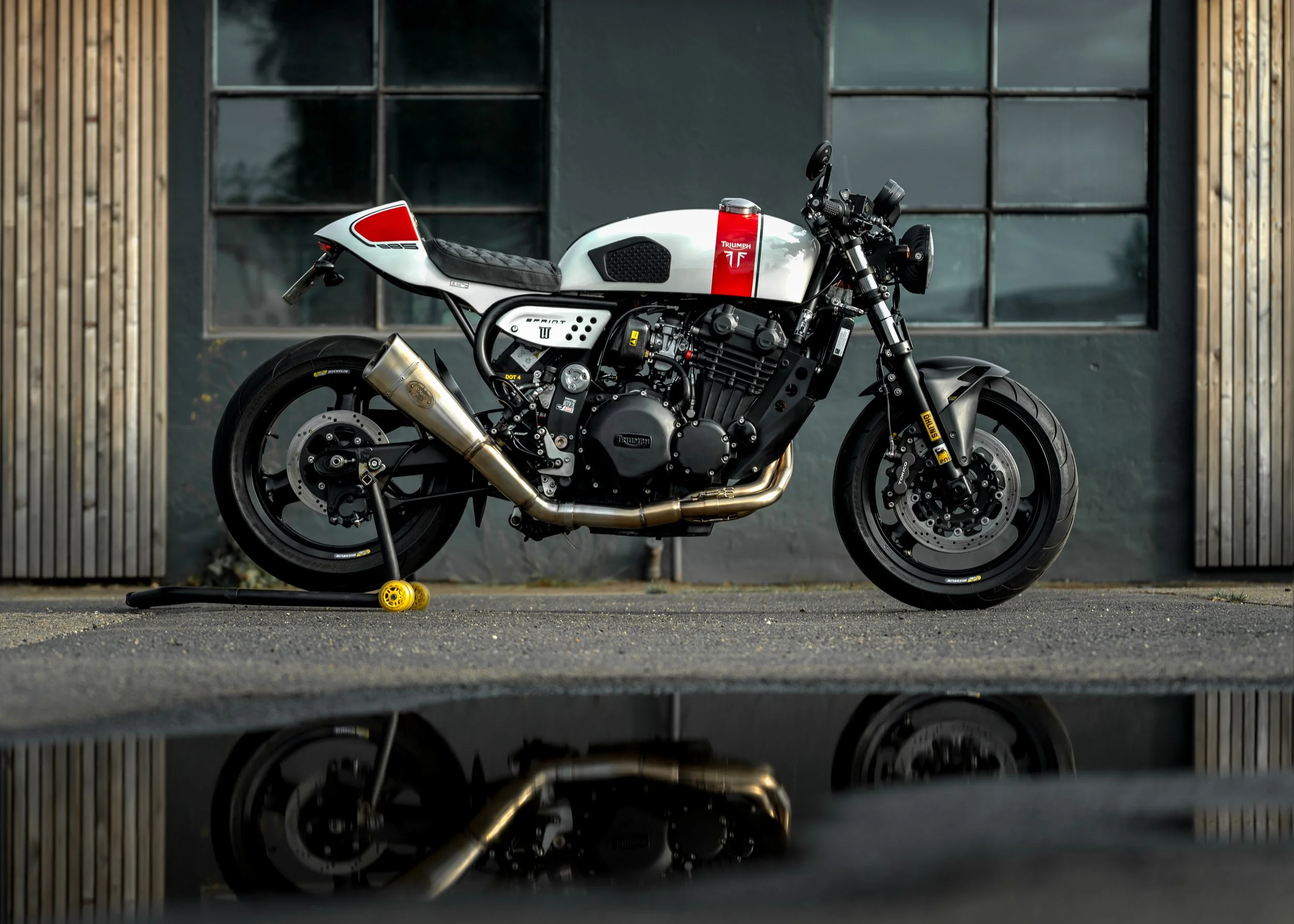 A black and white Triumph motorcycle with a custom seat, parked on a surface with its reflection visible in a puddle. The background features a building with wooden and metal paneling and large windows.