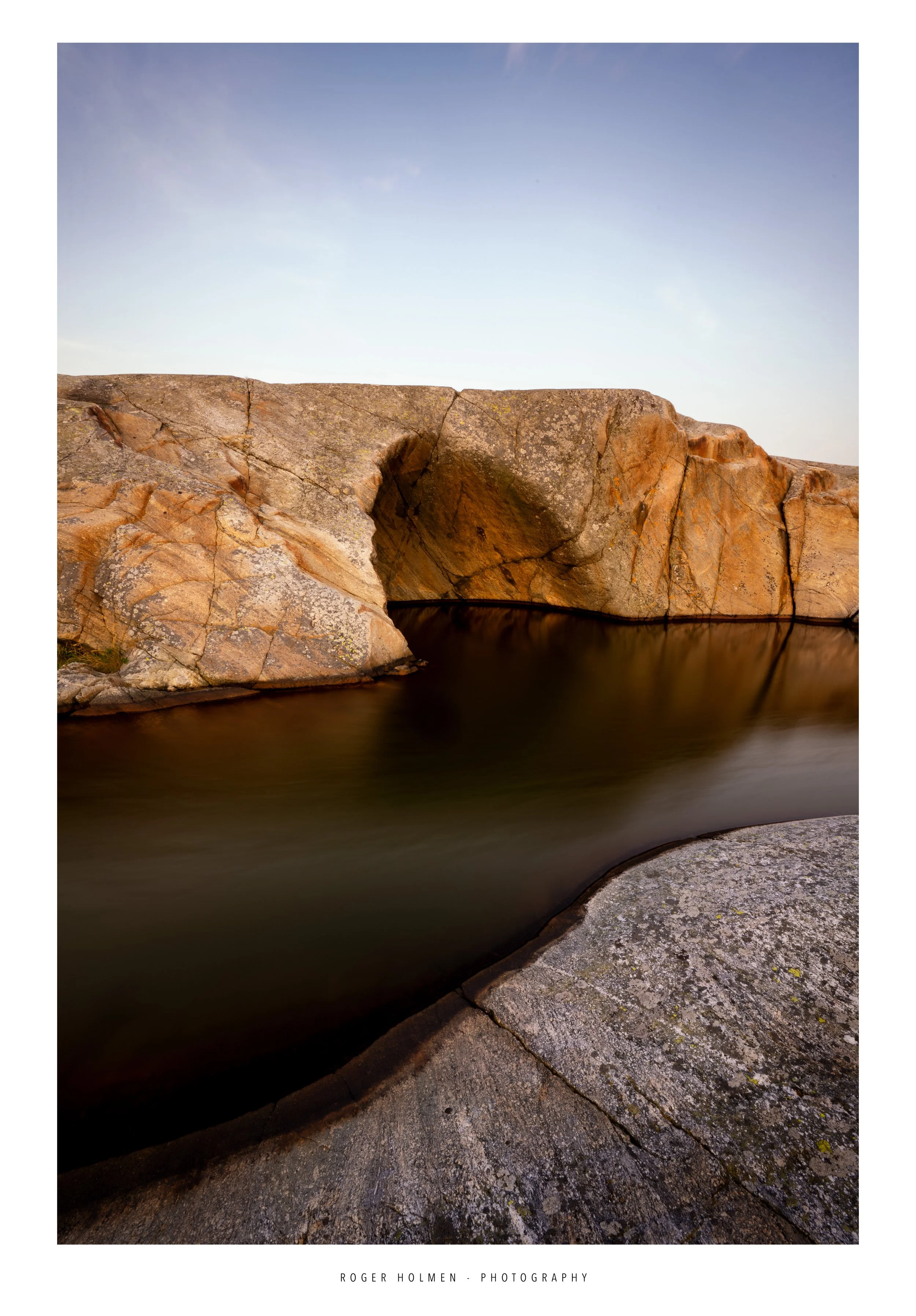 Large rocky formation with a small cave opening along a calm body of water, under a clear sky.