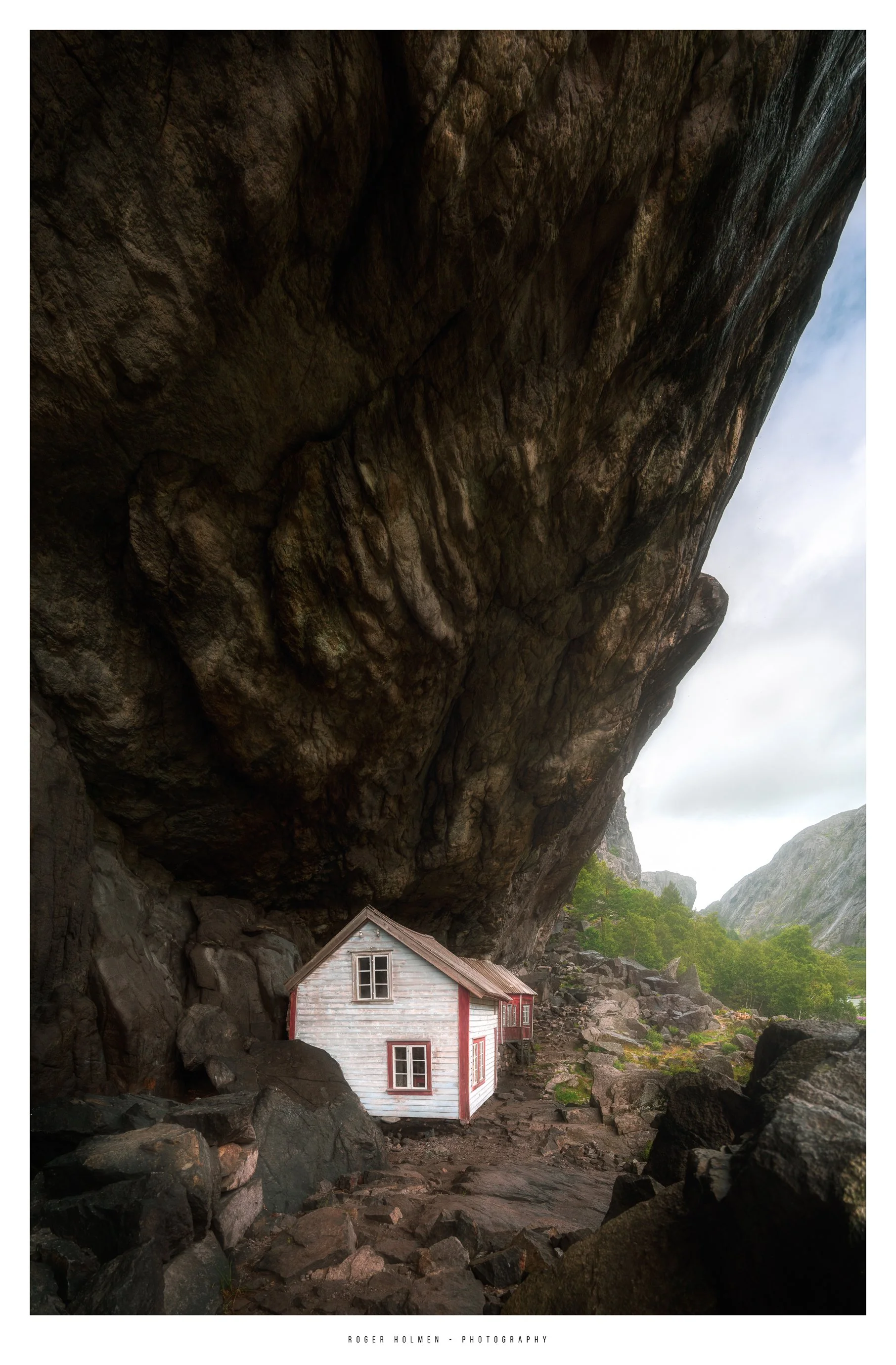 A small, white wooden house with red trim located underneath a large, overhanging rock formation in a rocky mountain landscape with green trees and cloudy sky.