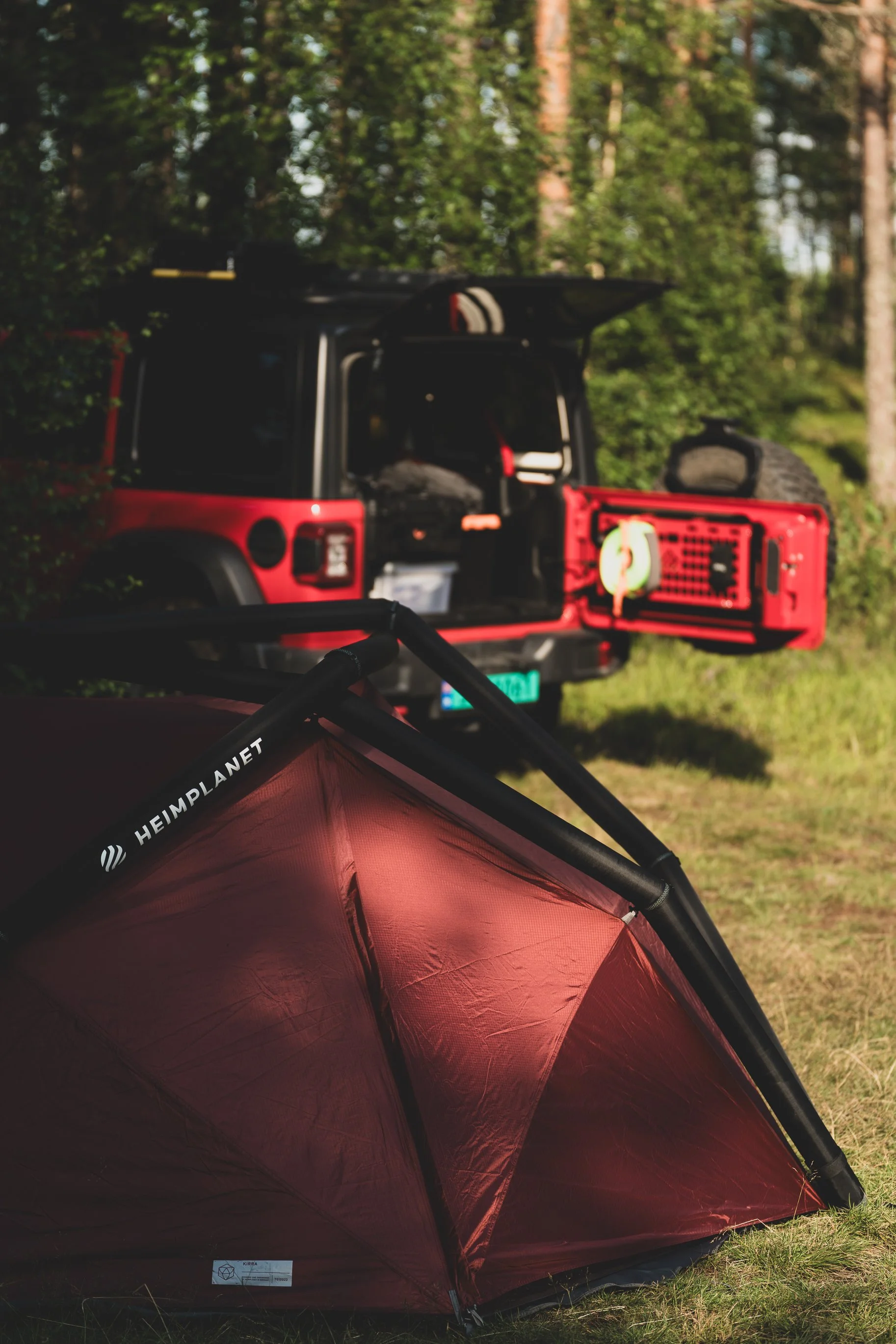 A red camping tent labeled 'HEIMPLANET' set up on grassy ground near a red off-road vehicle with its back hatch open, showing camping gear inside, in a wooded area.