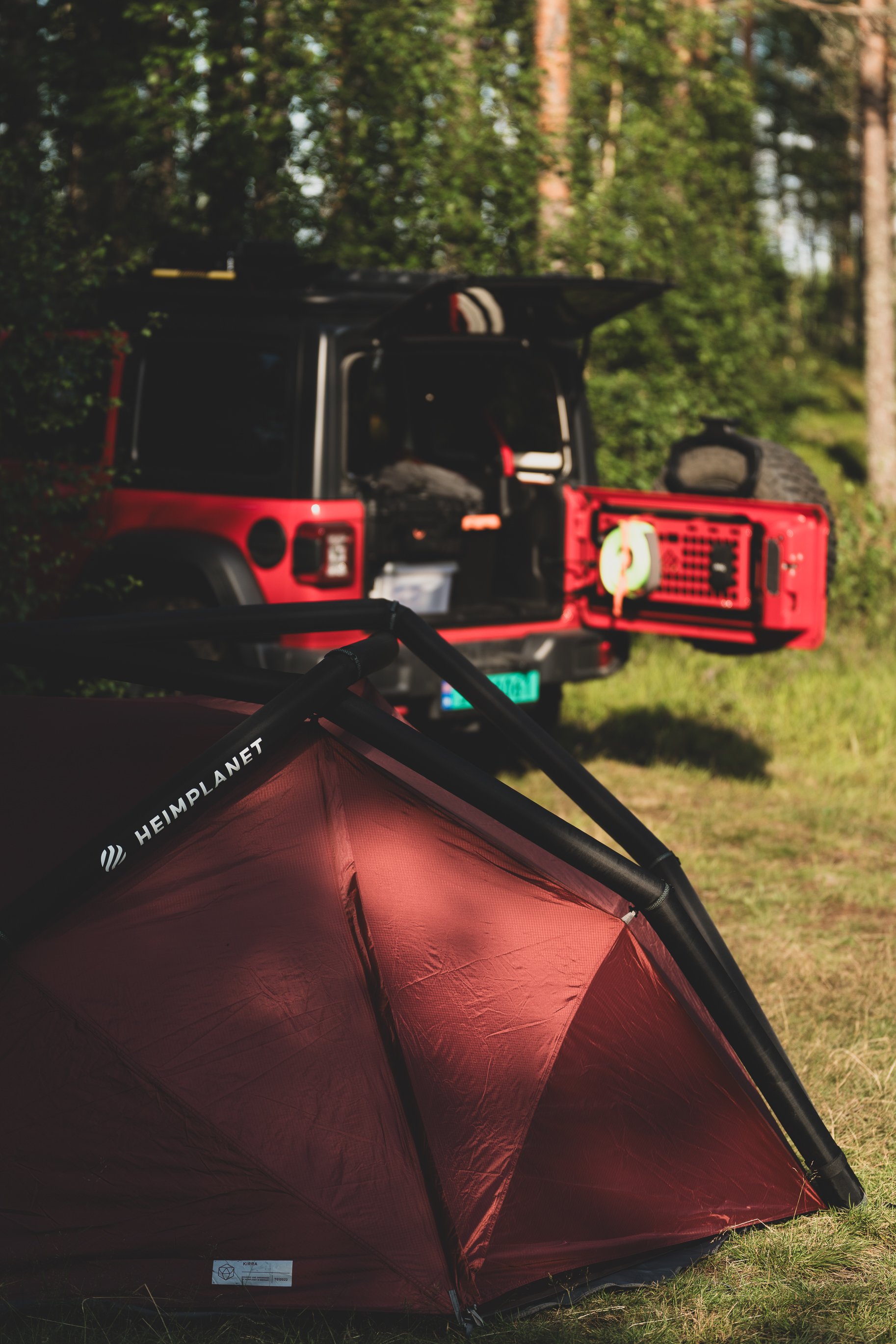 A red camping tent labeled 'HEIMPLANET' set up on grassy ground near a red off-road vehicle with its back hatch open, showing camping gear inside, in a wooded area.