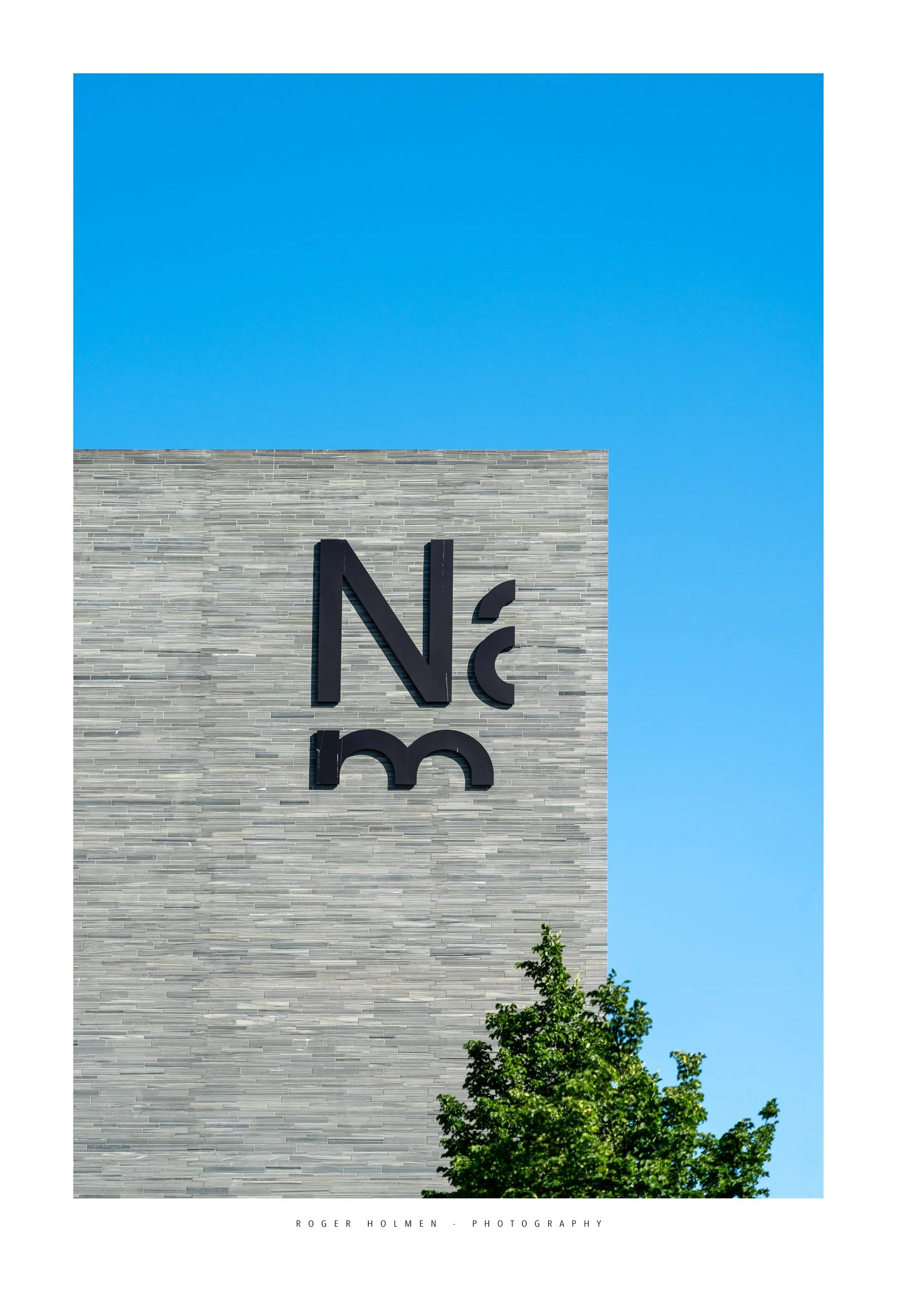 Part of a modern building with a gray brick exterior, displaying the letters 'N' and 'c' with a stylized design, against a bright blue sky, and a green leafy tree in the lower right corner. Arkitektur foto oslo. Nasjonal Museet.
