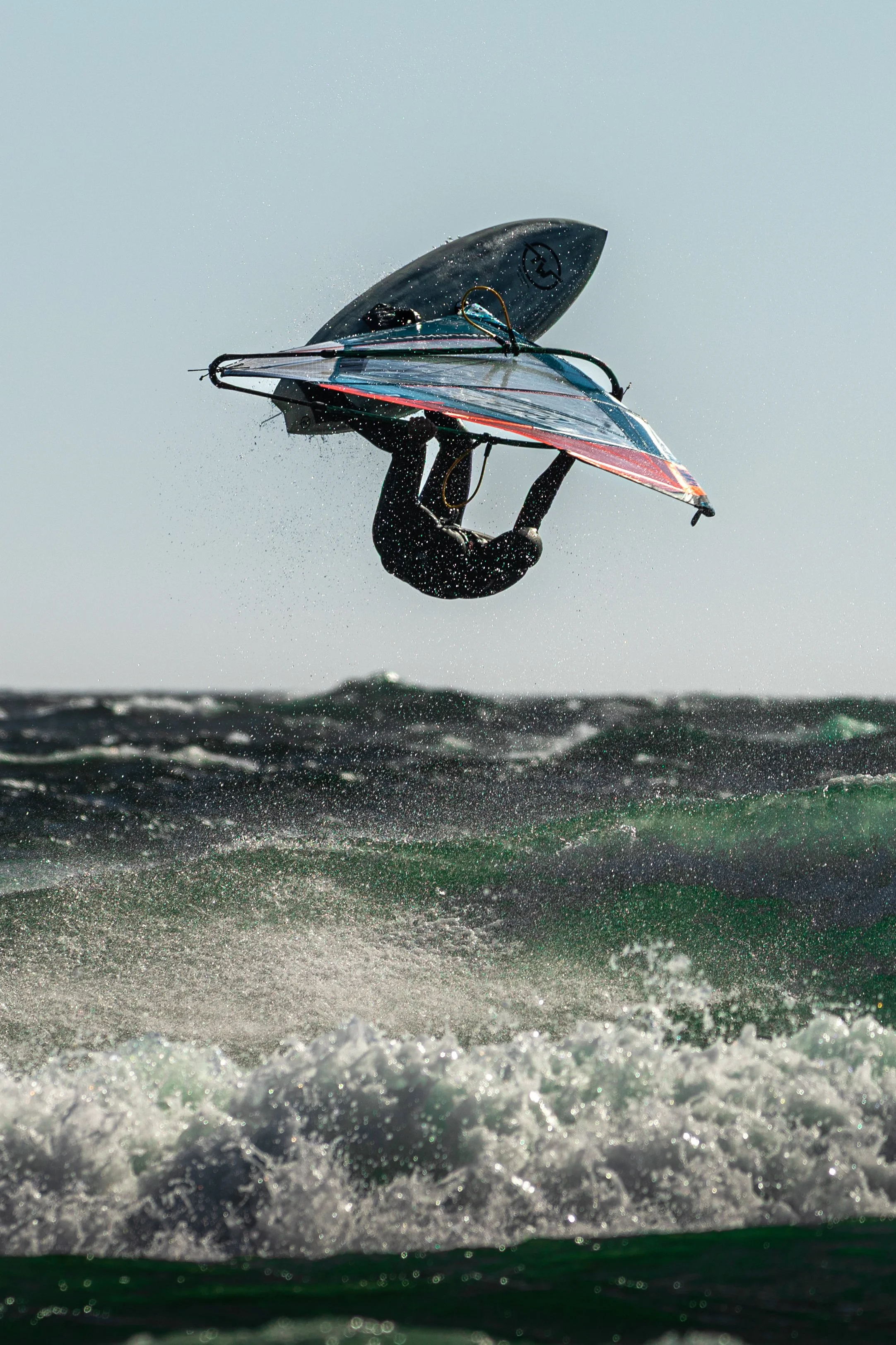 A person wearing a black wetsuit windsurfing on choppy water with a large sail and a black surfboard, mid-air above waves. Location, Saltstein, Norway.