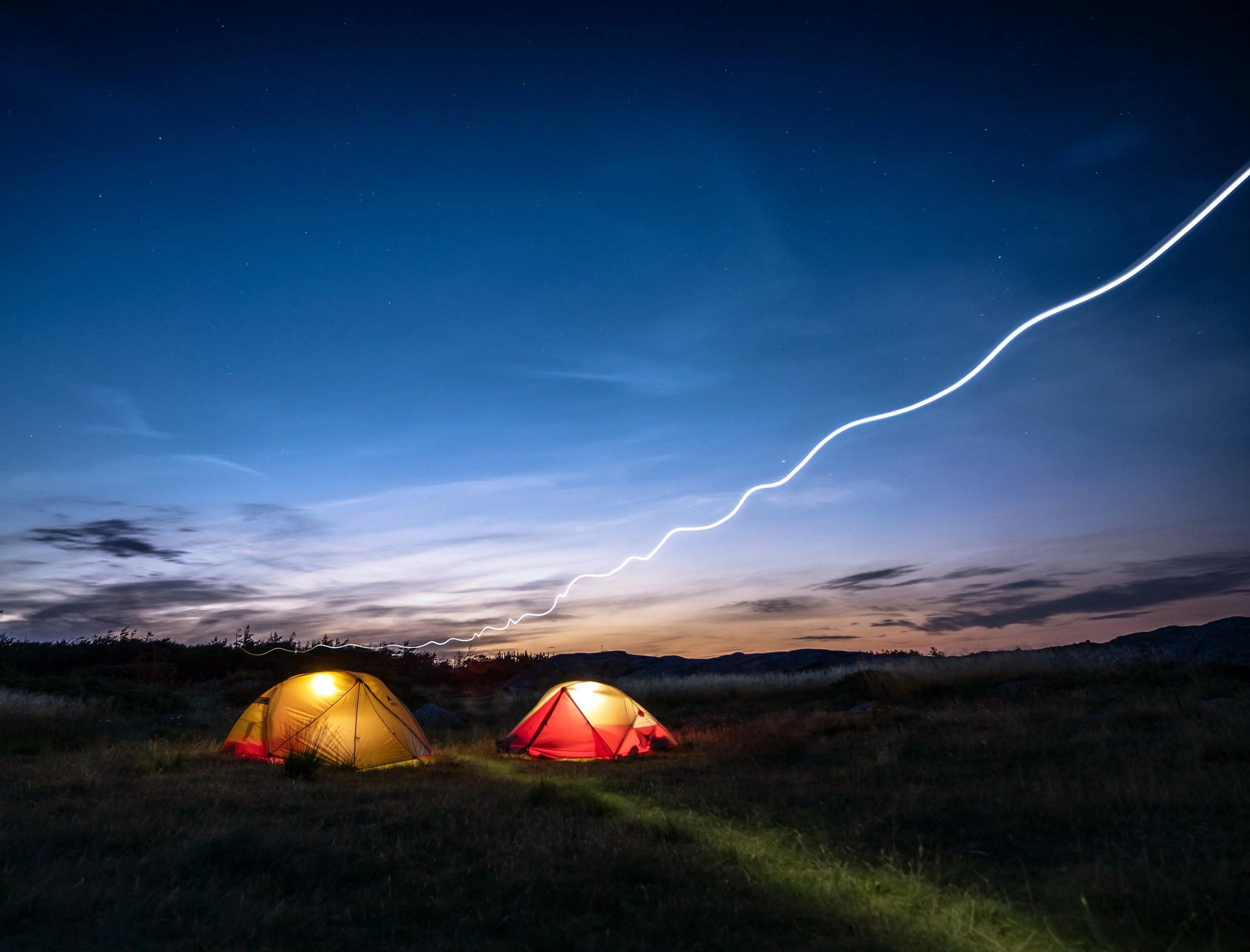 Nighttime scene of two tents (yellow and red) on a grassy field with a dark sky, streaks of lightning visible in the sky, and distant mountains.