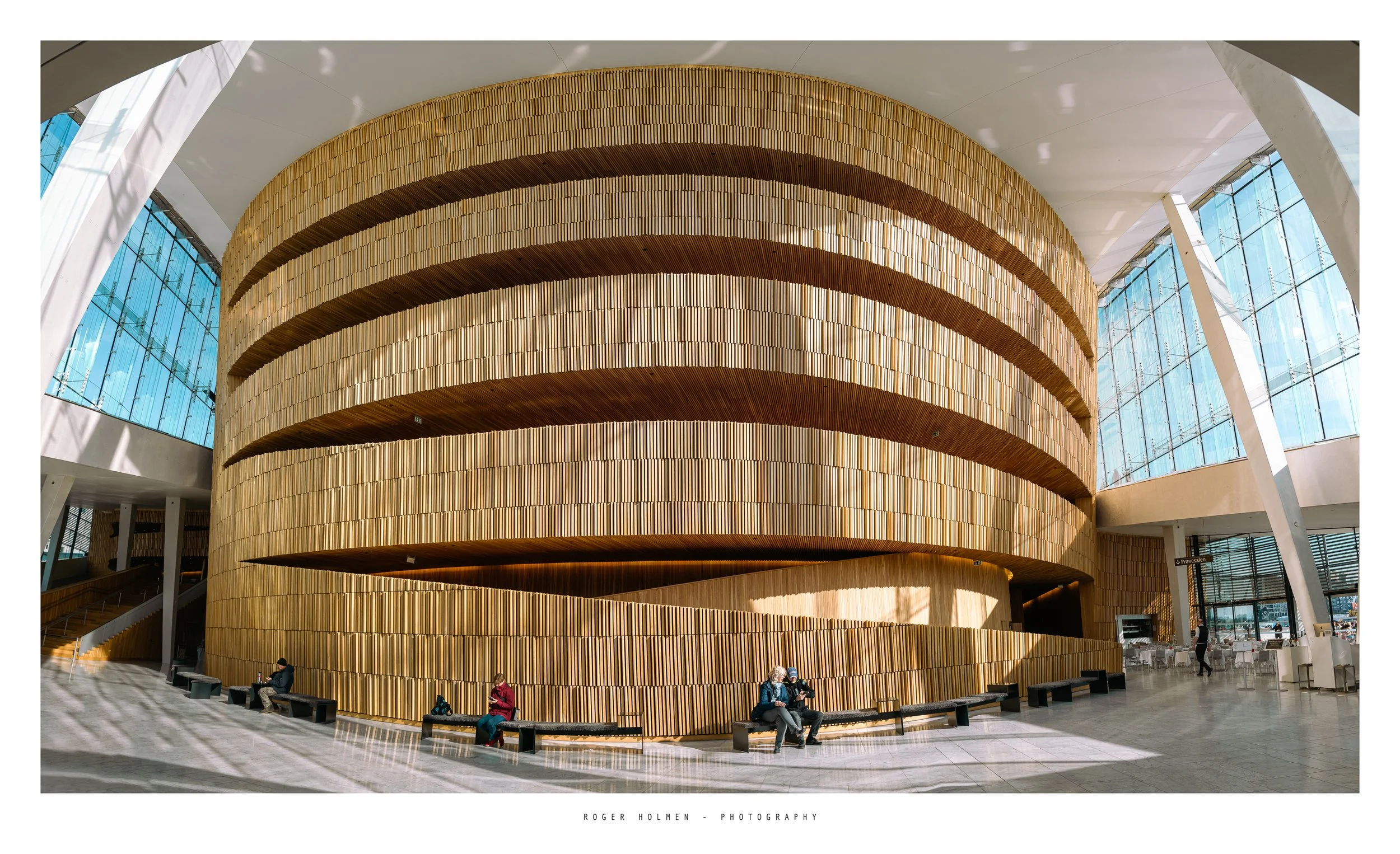 Interior of a modern building with a large, curved, multi-level wooden structure, glass walls, and several people sitting and walking inside.