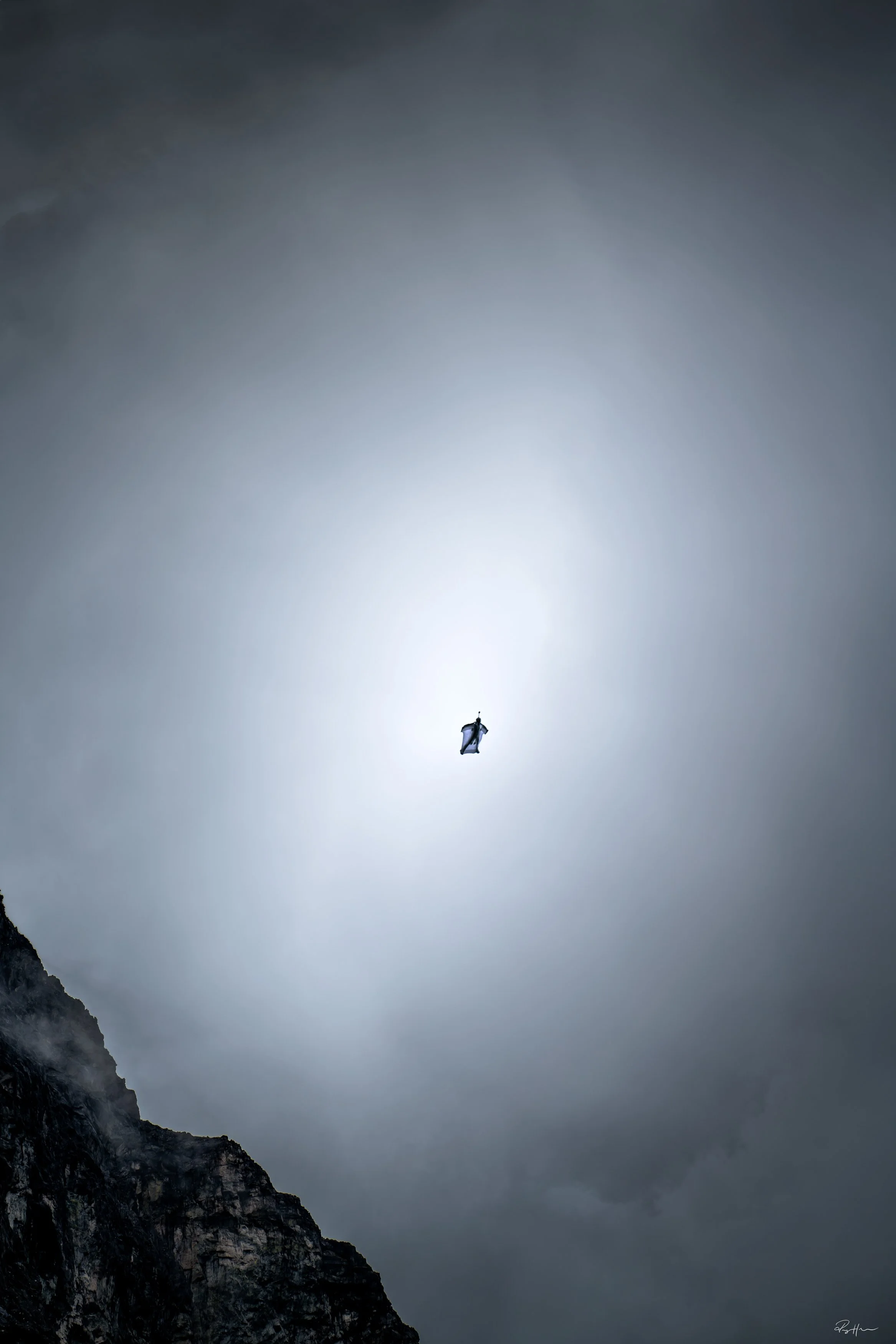 Wingsuit flight in a cloudy sky above a mountain landscape.