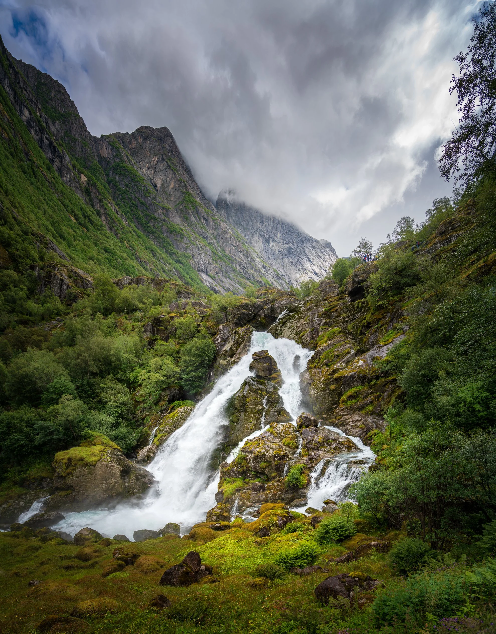 A waterfall cascading down rocky terrain surrounded by lush green trees and moss, with towering mountains in the background under a cloudy sky and a group of people walking on a trail on the right side.