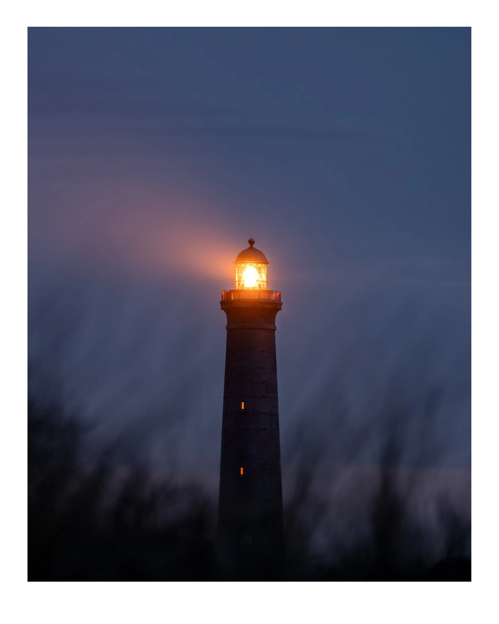 Lighthouse at dusk with its light shining brightly into the evening sky.