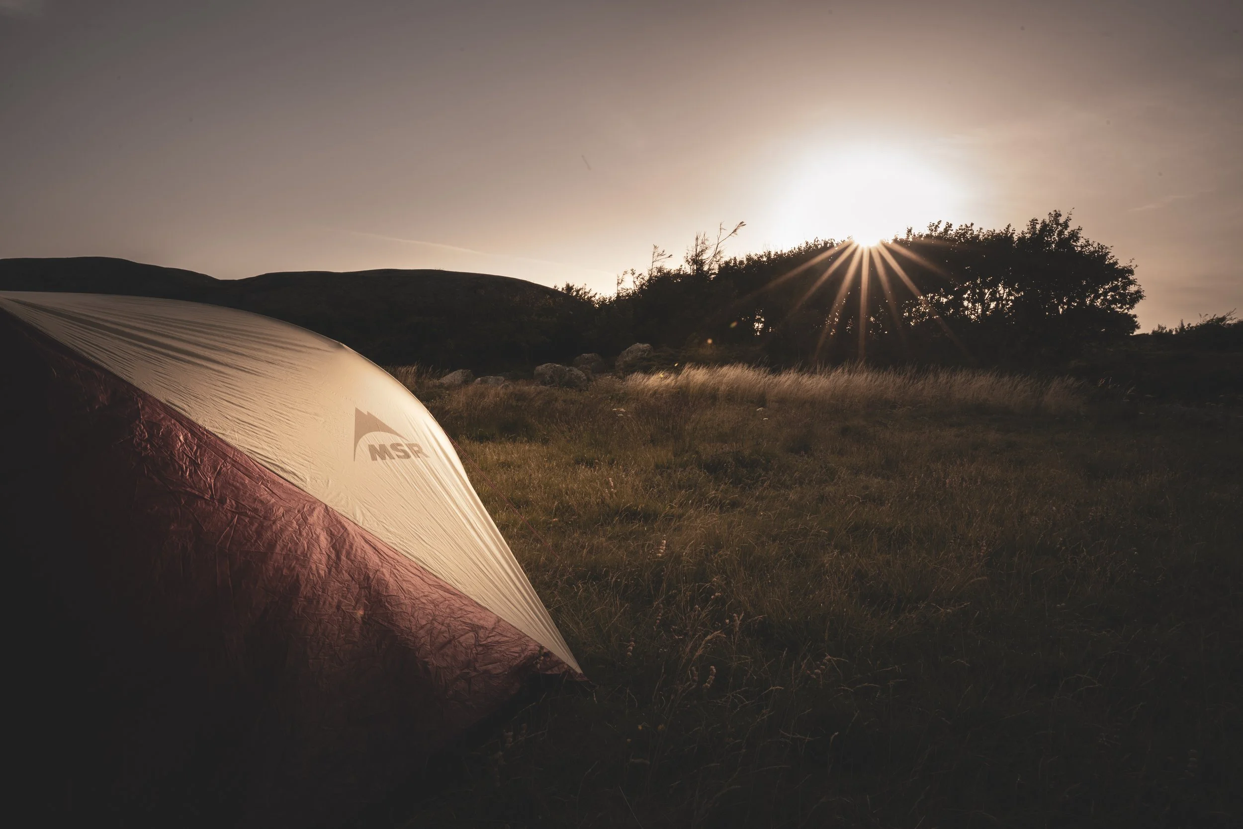 A campsite with a tent in a grassy field at sunset, trees and hills in the background. MSR Tents.
