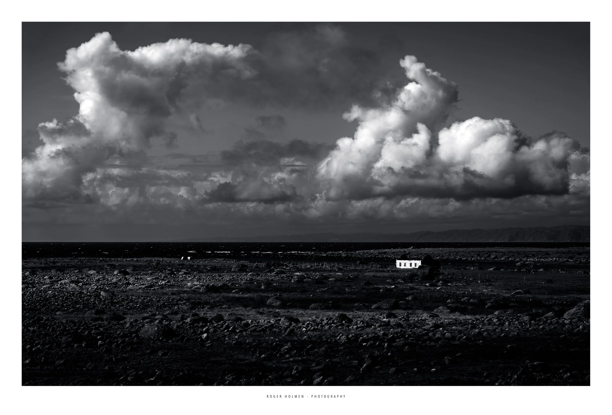 A black and white photograph of a vast, rocky landscape with a small, white building in the distance near the horizon. The sky is filled with large, dramatic clouds.
