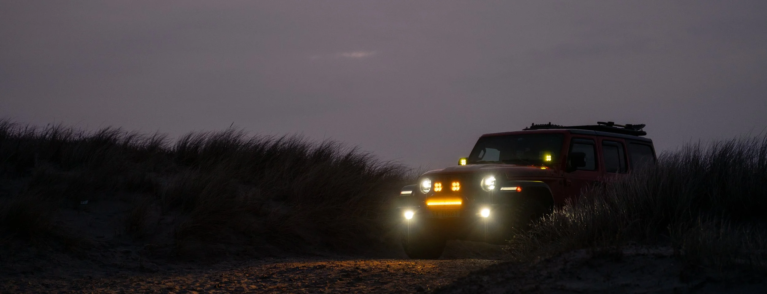 A red Jeep with yellow headlights and additional off-road lights is parked on sandy dunes at dusk, surrounded by tall grass.