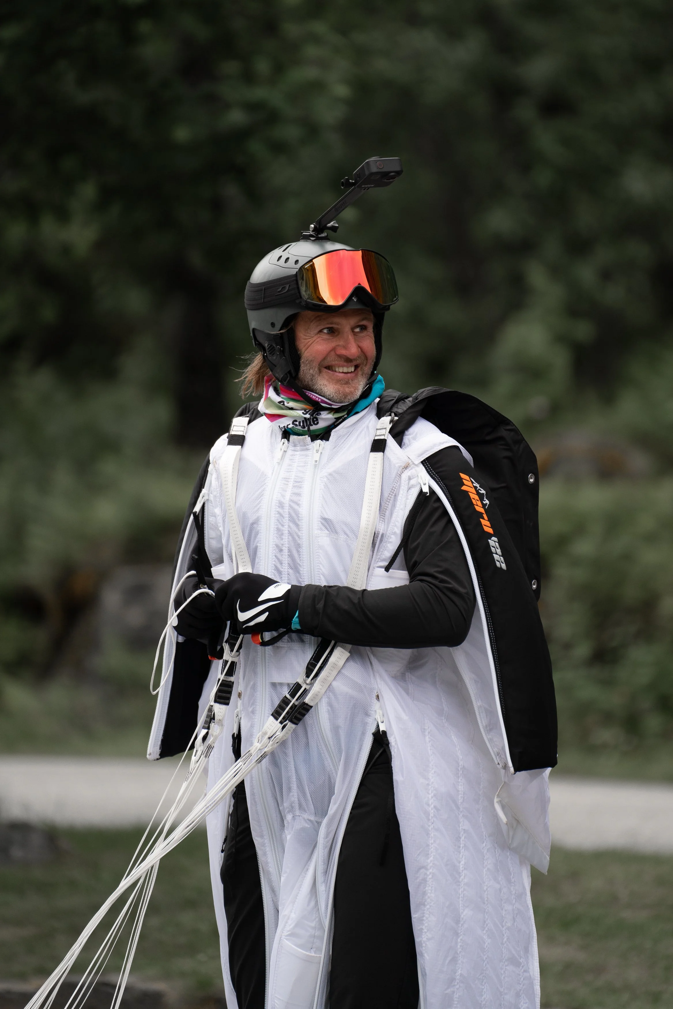 A man smiling outdoors, wearing a white wingsuit, a helmet with a camera, and gloves, preparing for a parachute jump.