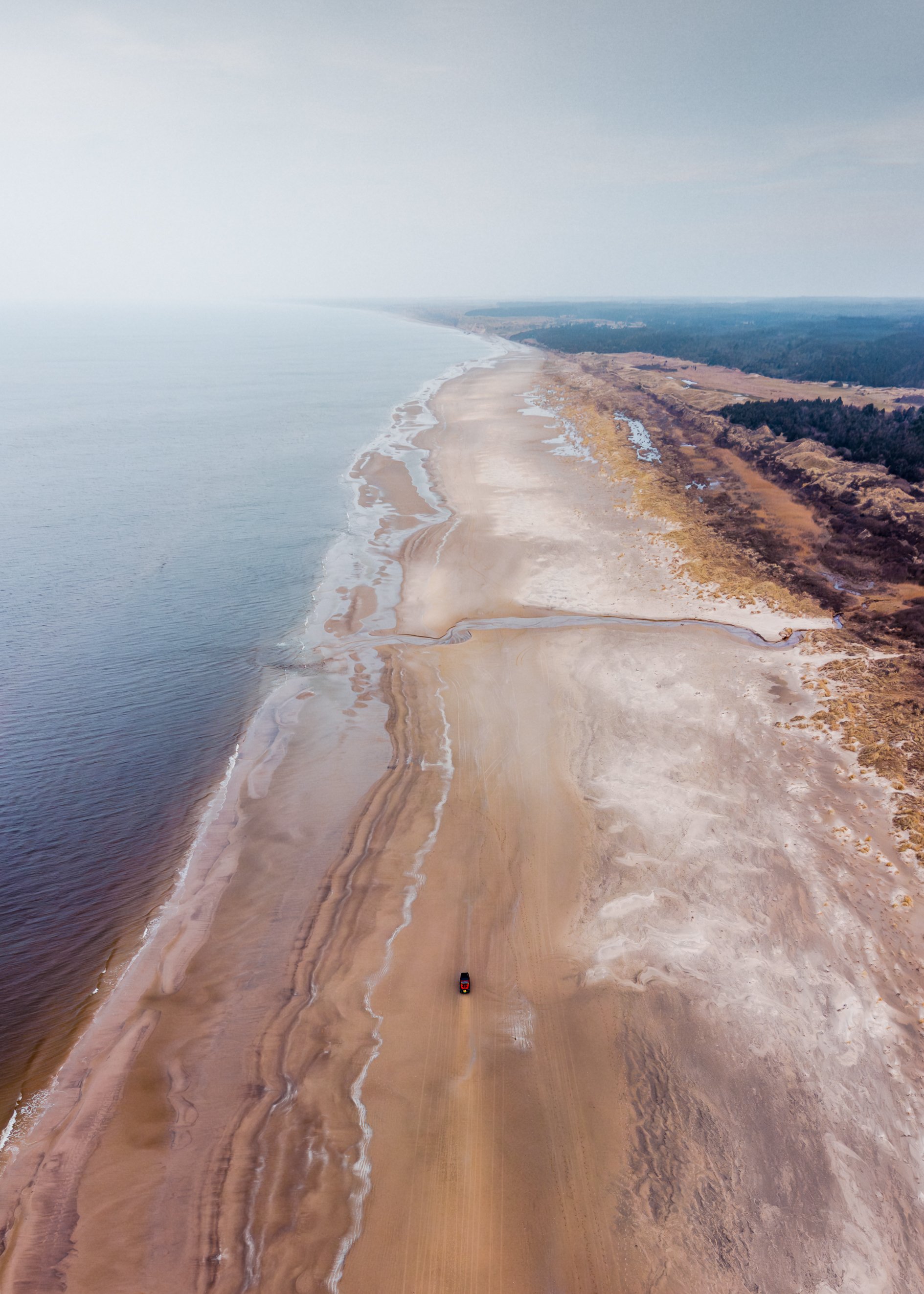 Aerial view of a long, sandy beach with waves on the left and land on the right, including a car on the beach.