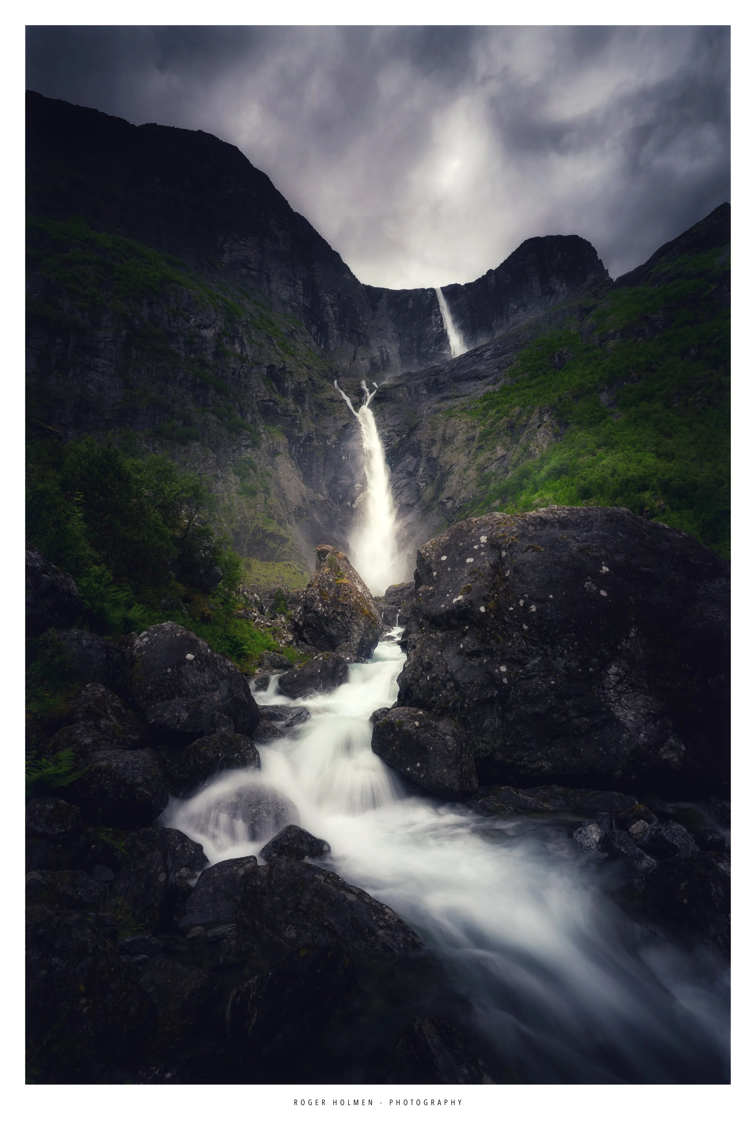 A multi-tiered waterfall flowing down a mountain surrounded by green trees and large rocks under a cloudy sky. Mardalsfossen.