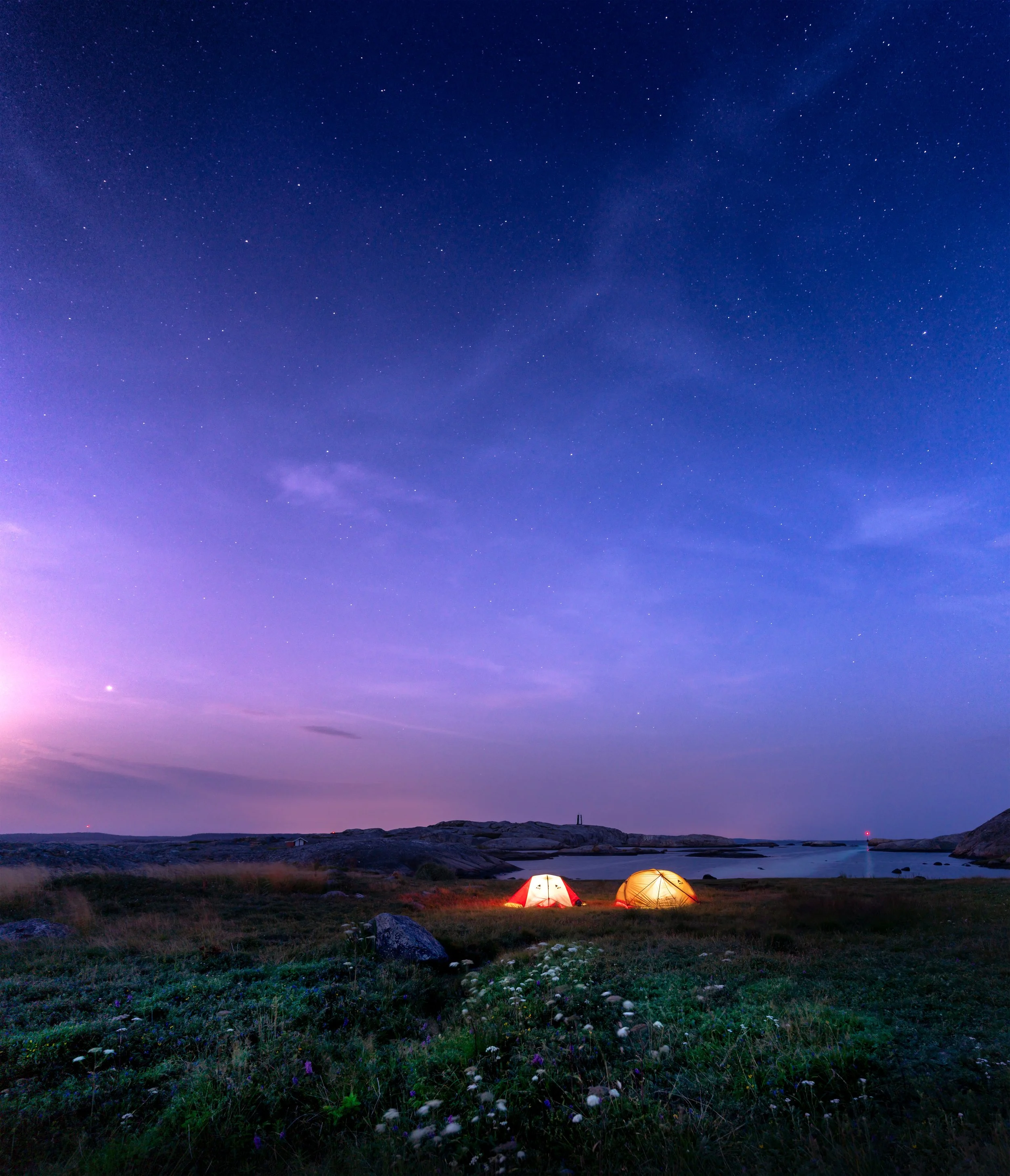 Nighttime scene with two illuminated tents on a grassy shoreline near water, under a star-filled sky with purple and blue hues.