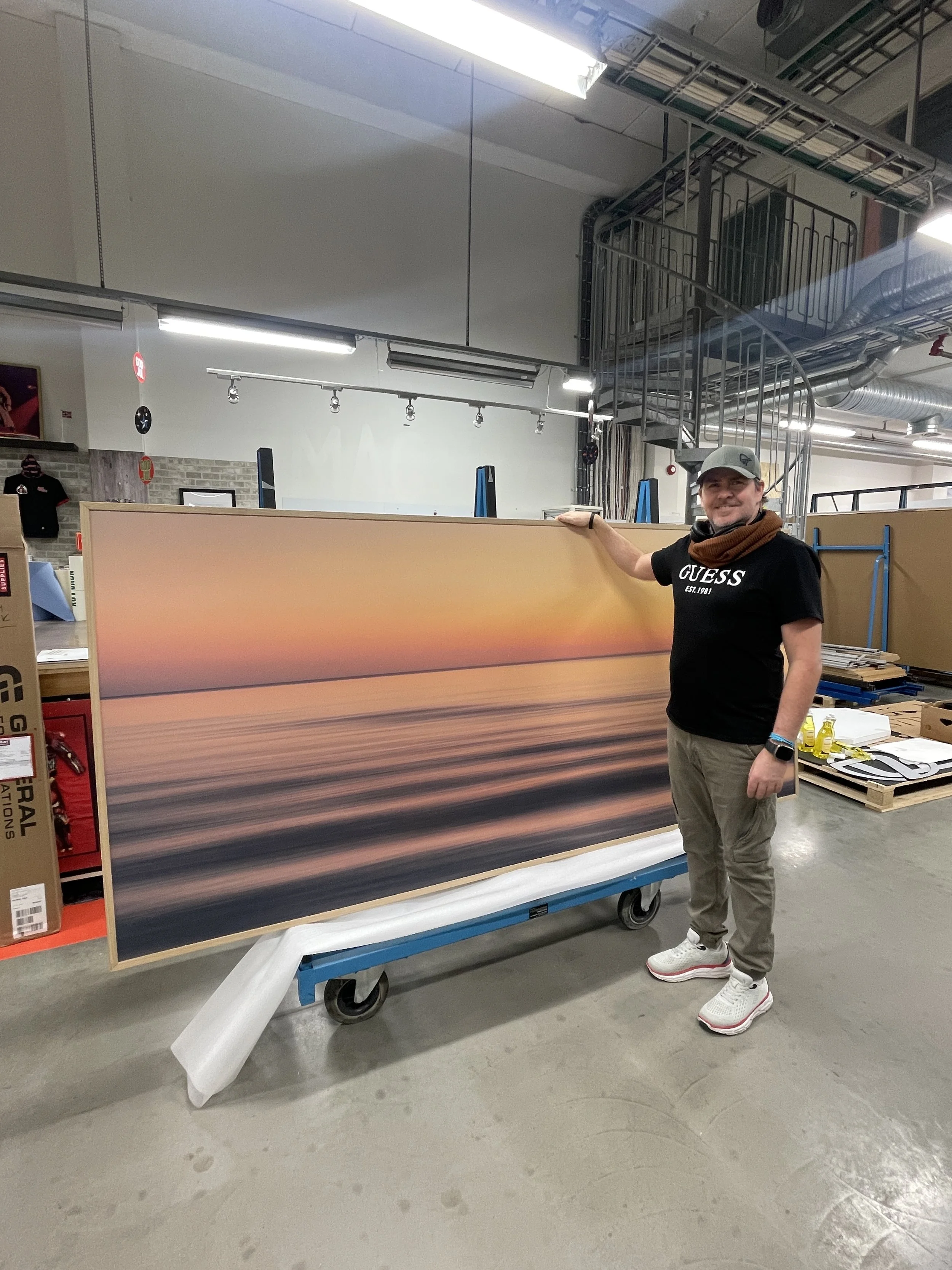 A man wearing a Guess T-shirt and a baseball cap stands next to a large framed photograph of a sunset over water, placed on a cart in an industrial or warehouse setting.
