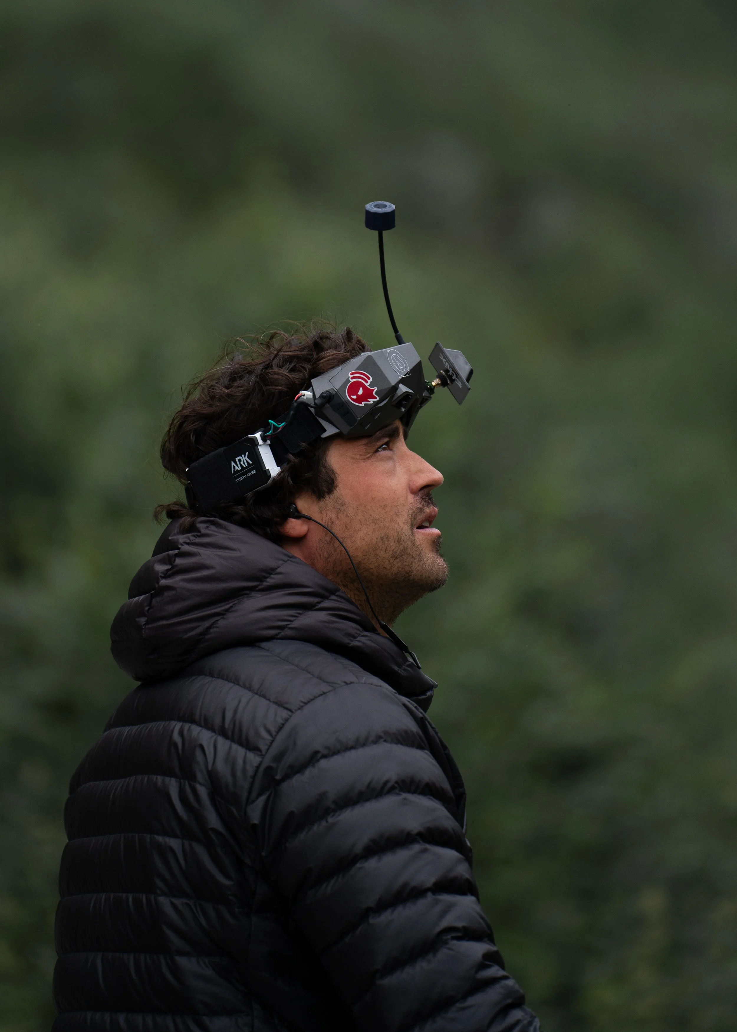 Man wearing a black puffer jacket and a head-mounted drone controller outdoors, looking thoughtfully into the distance.