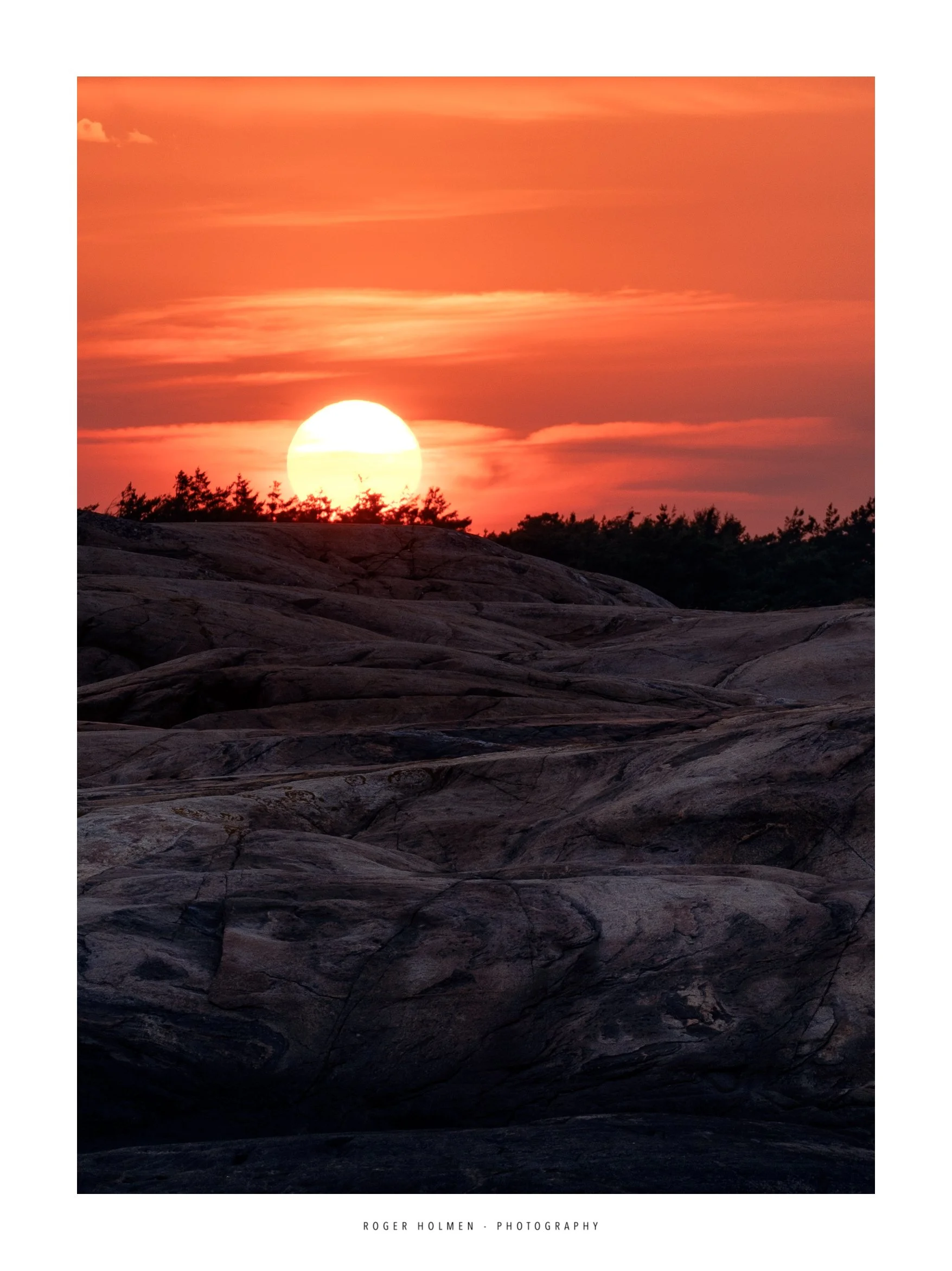 Photo of a sunset with the sun partially visible touching the horizon, with pink and orange sky and some clouds, above dark rocks and silhouetted trees. Location Herføl.