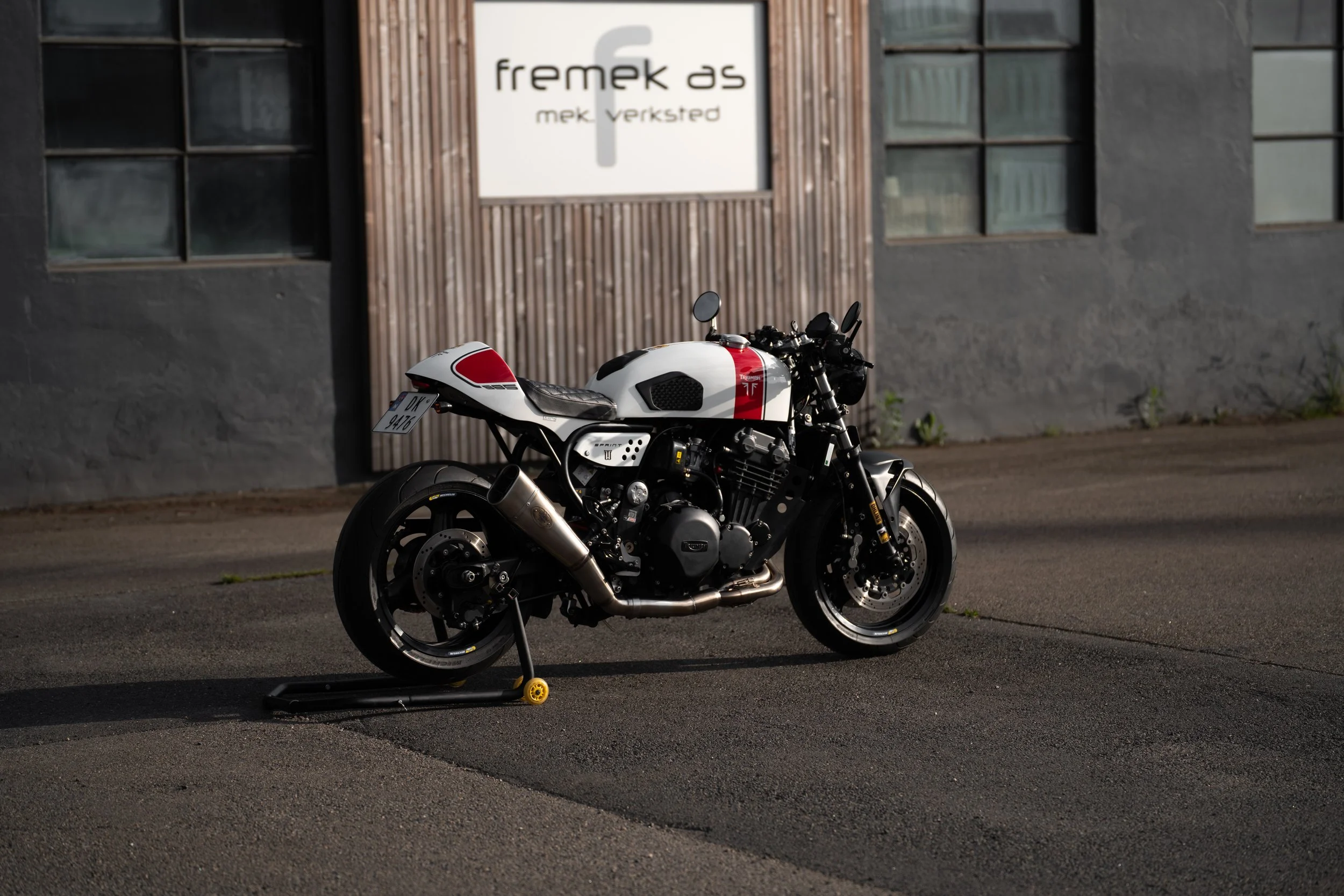 A black and white motorcycle with red accents parked on asphalt in front of a gray building with a wooden accent wall and a sign that reads 'fremak as mek verksted'.